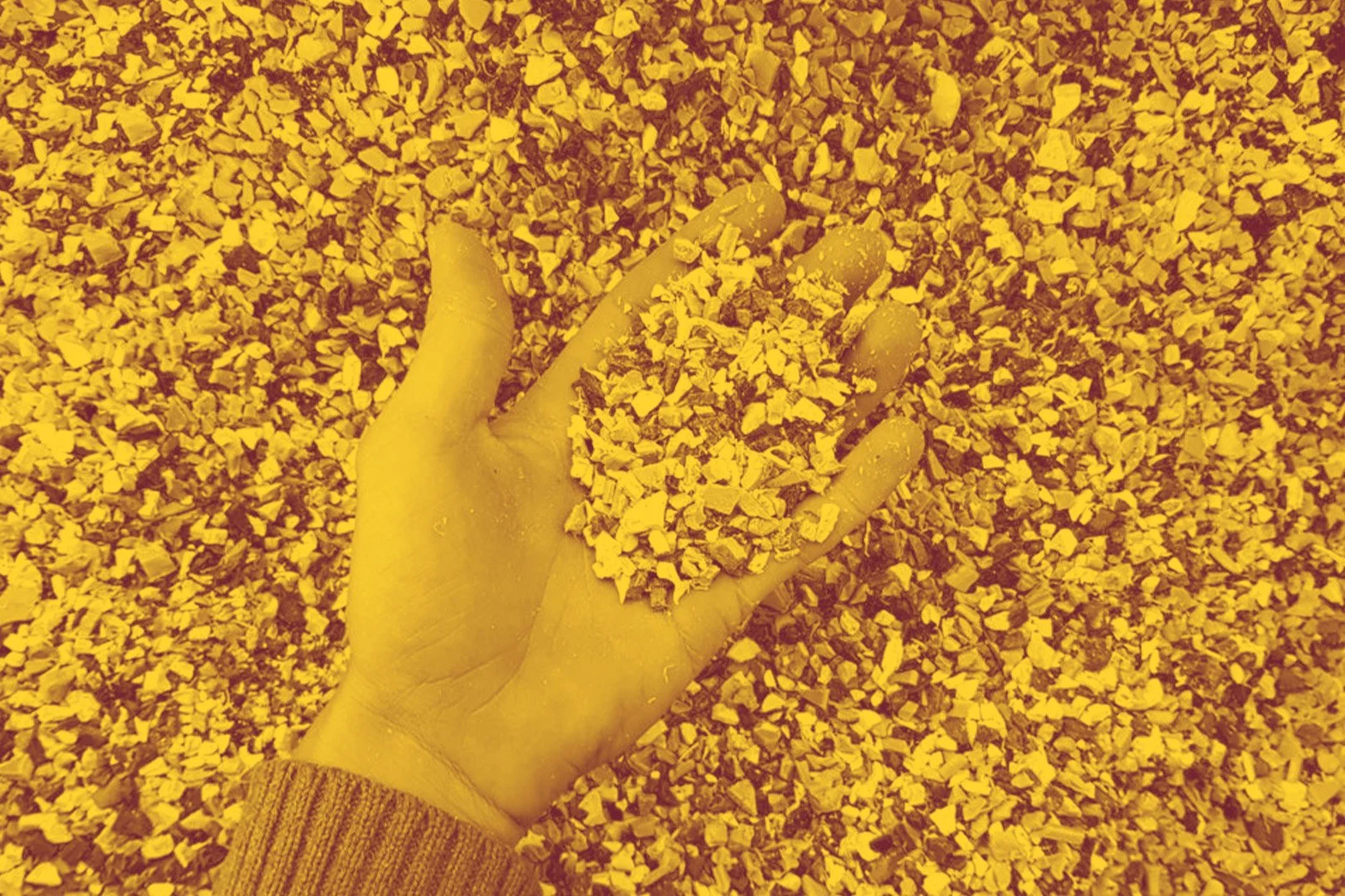 A person wearing a yellow glove holding a handful of small yellowish-brown crushed recycled plastic.