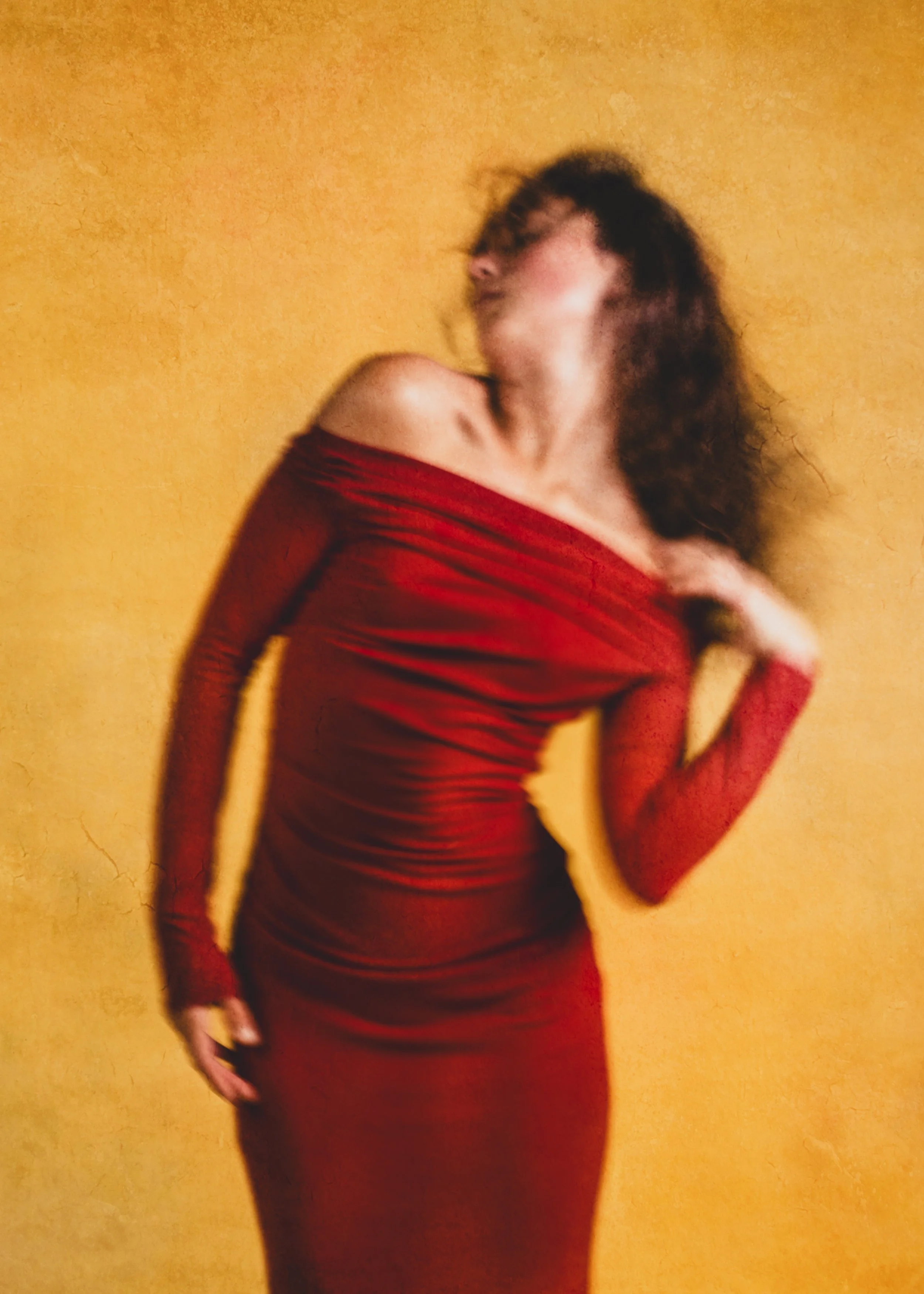 A woman with wavy hair dancing, wearing a red off-the-shoulder dress, posing against a yellow background.