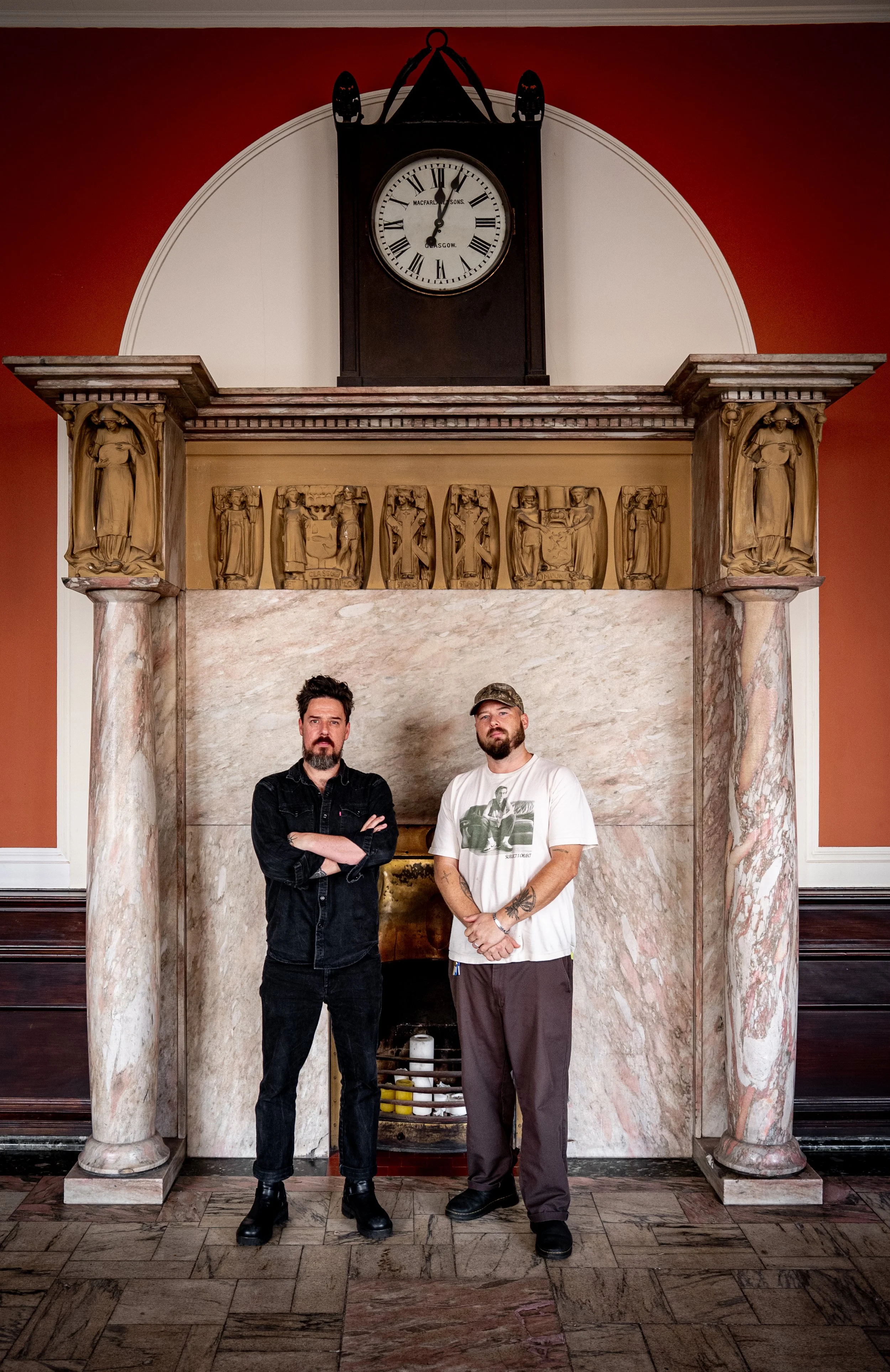 Two men standing in front of a decorative marble fireplace with a wooden mantle and ornate carvings, in a room with red walls and a large clock hanging above.