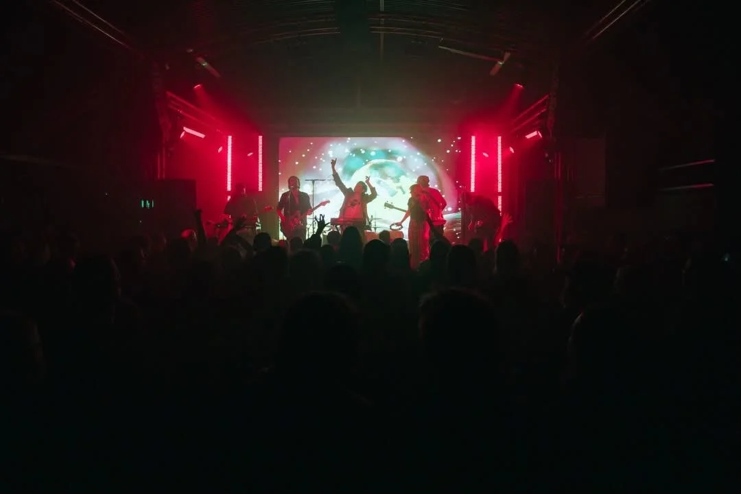 A live band performs on stage with vibrant red and blue lighting, an animated backdrop, and an audience watching in the dark.