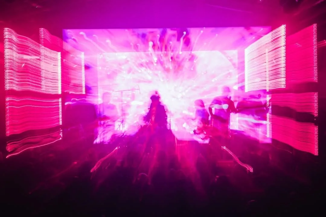 Blurred image of a live concert with vibrant pink and purple lights, silhouettes of musicians on stage, and motion effects.