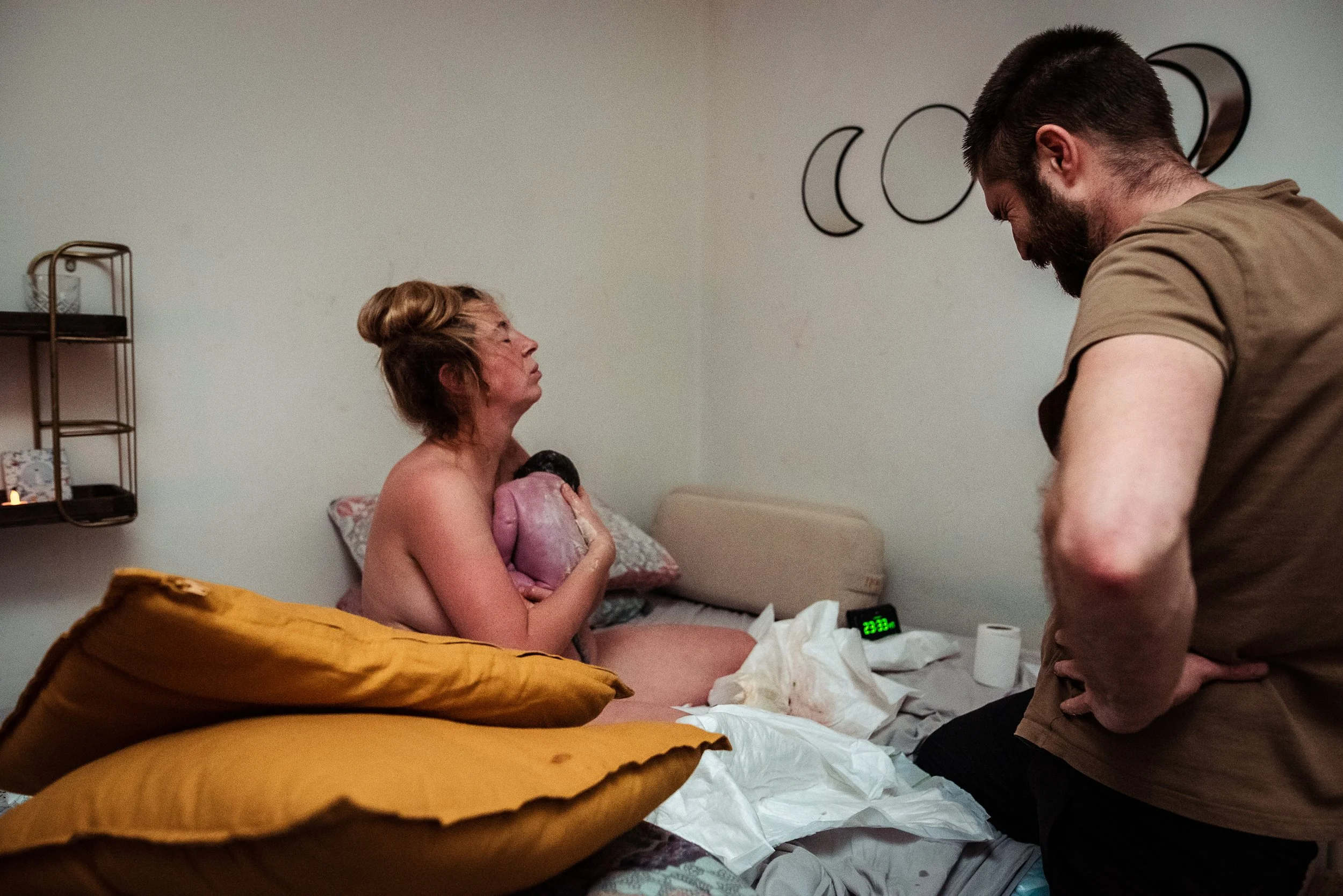 A woman gives birth in a hospital room, holding her newborn baby to her chest, with a man standing nearby looking down at her.