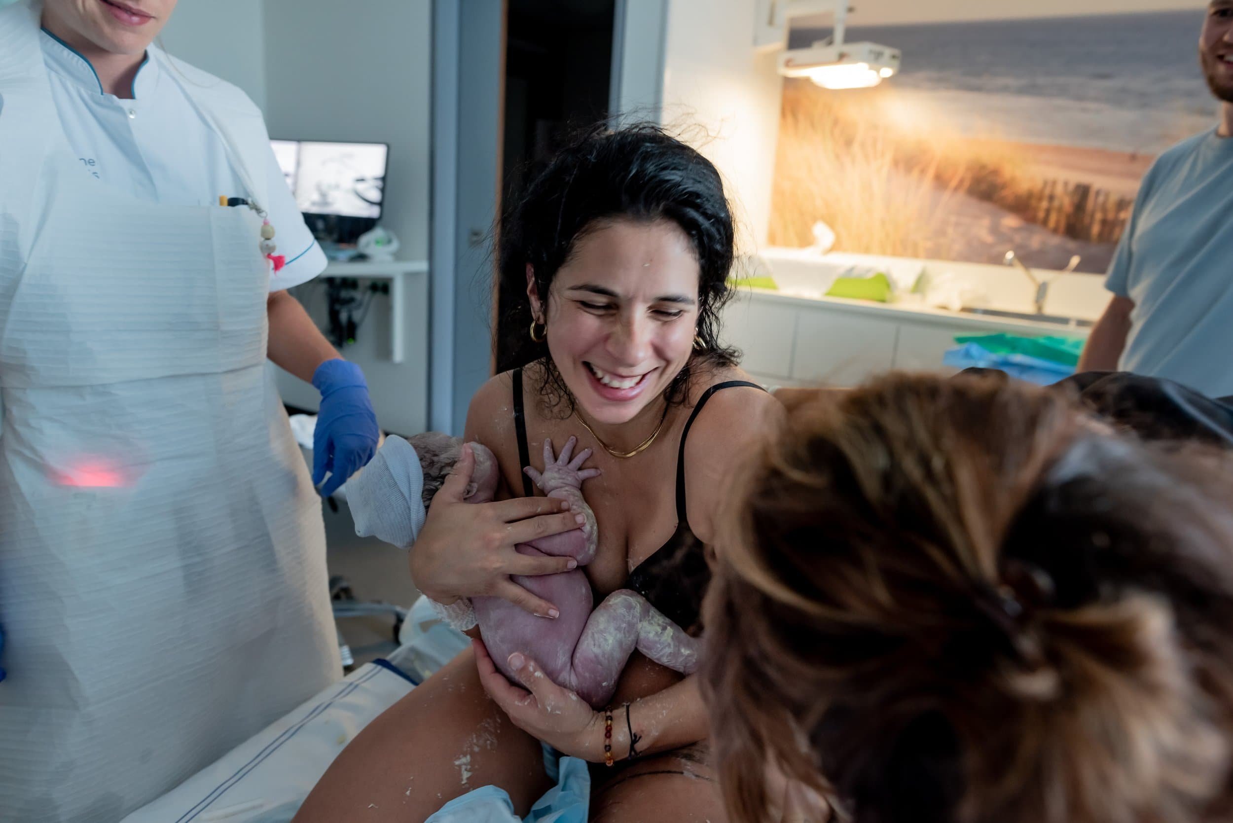 A woman celebrates giving birth in a hospital room, holding a newborn baby with assistance from medical staff.