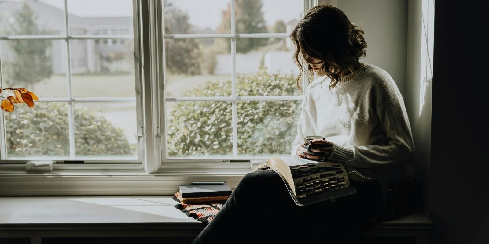 Woman with curly hair sitting in a window seat reading a book and holding a mug, soft natural light — recovering from ADHD burnout in motherhood
