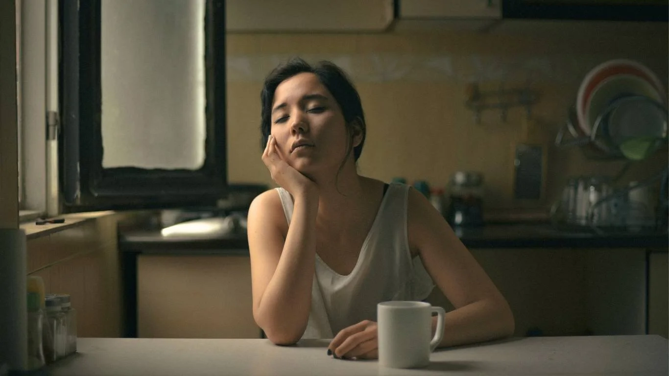A woman sitting alone at a kitchen table with a mug, hand resting against her face — the quiet depletion of setting limits as an ADHD mum