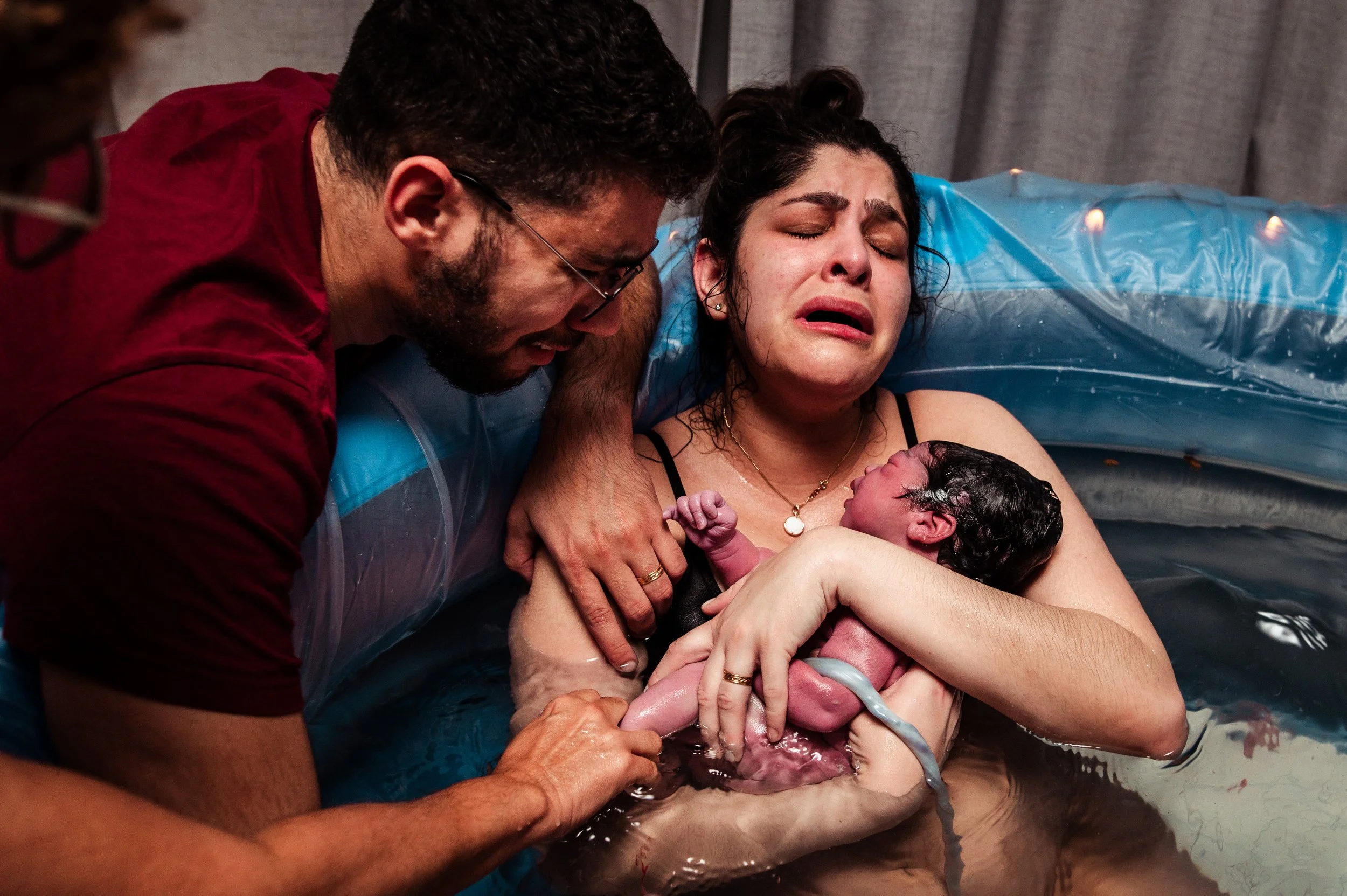 A woman giving birth in a birthing tub, crying with emotion as the baby is born and held by her, while a man supports her during the moment.