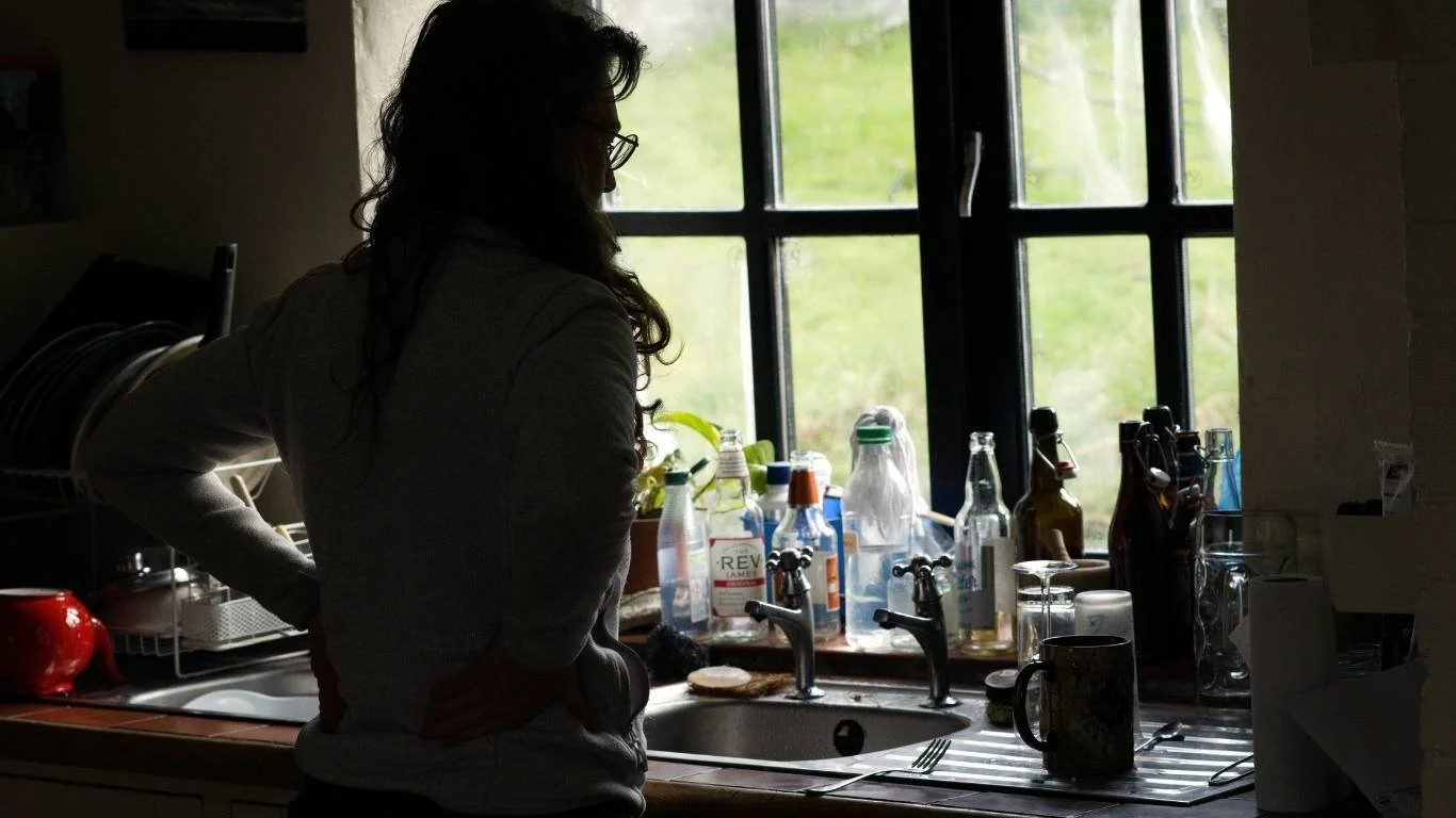 Woman standing in silhouette at a kitchen sink, looking out over a green field while surrounded by cluttered dishes and bottles, representing the quiet mental load women with late-diagnosed ADHD carry while masking.