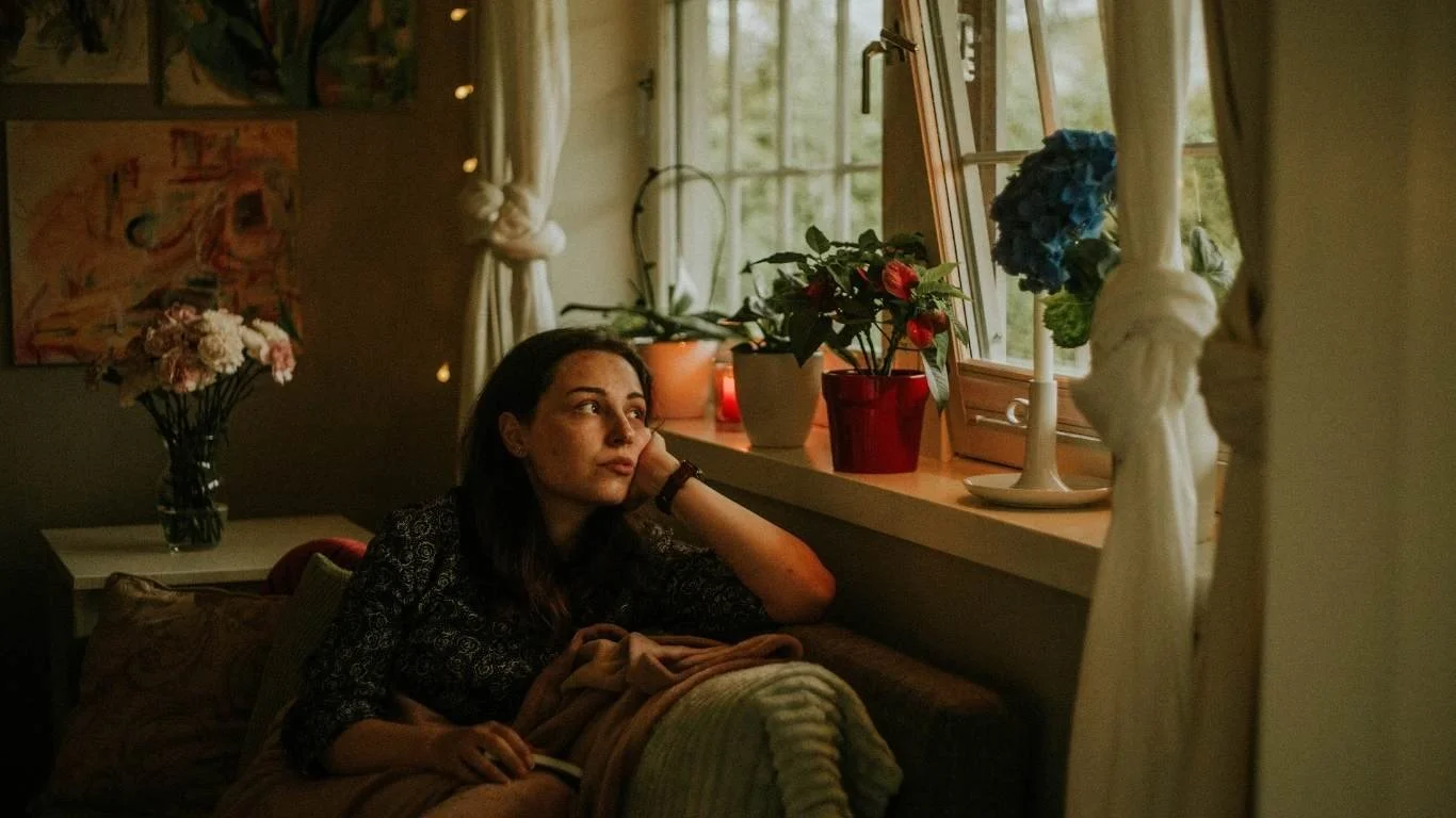 A mother sitting quietly by a window in soft daylight, on0e hand resting on face, taking a moment to steady herself