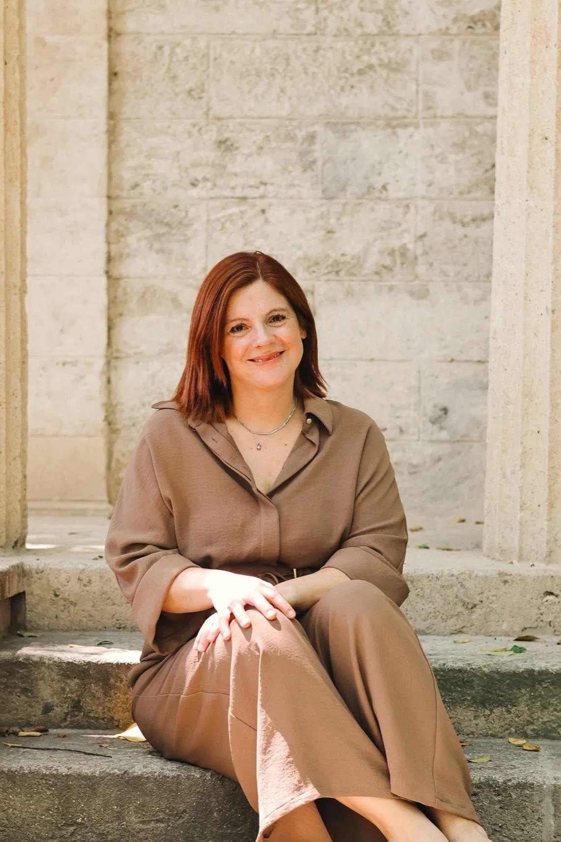 A woman with red hair sitting on stone steps outdoors, smiling, wearing a brown outfit, with a stone wall in the background.