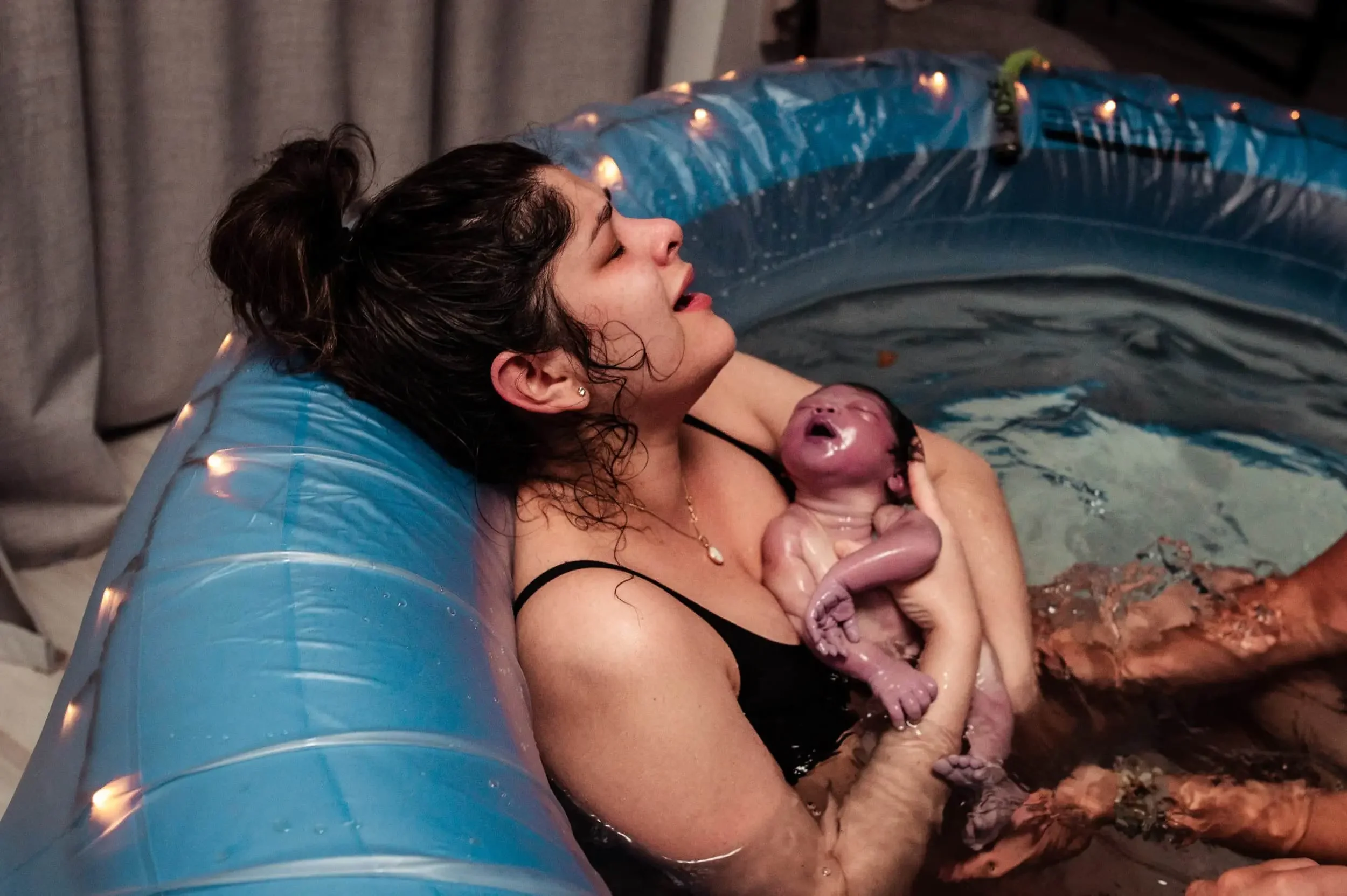 A woman giving birth in an inflatable birthing tub, holding a newborn baby in her arms, both appearing exhausted but joyful.