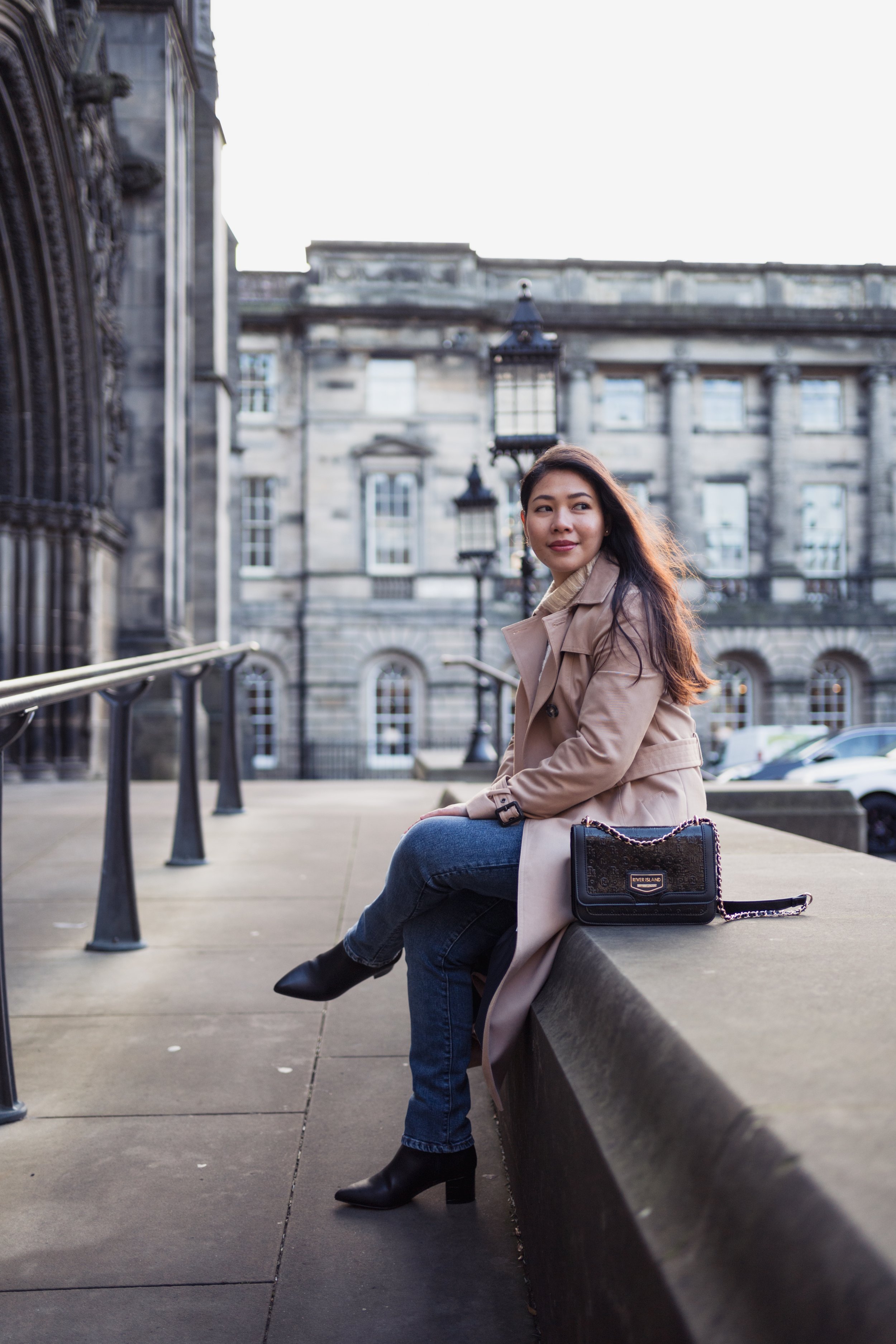 A woman with long dark hair, wearing a beige trench coat and black heeled boots, sitting on a stone ledge outside historic stone buildings with large windows and street lamps in the background.