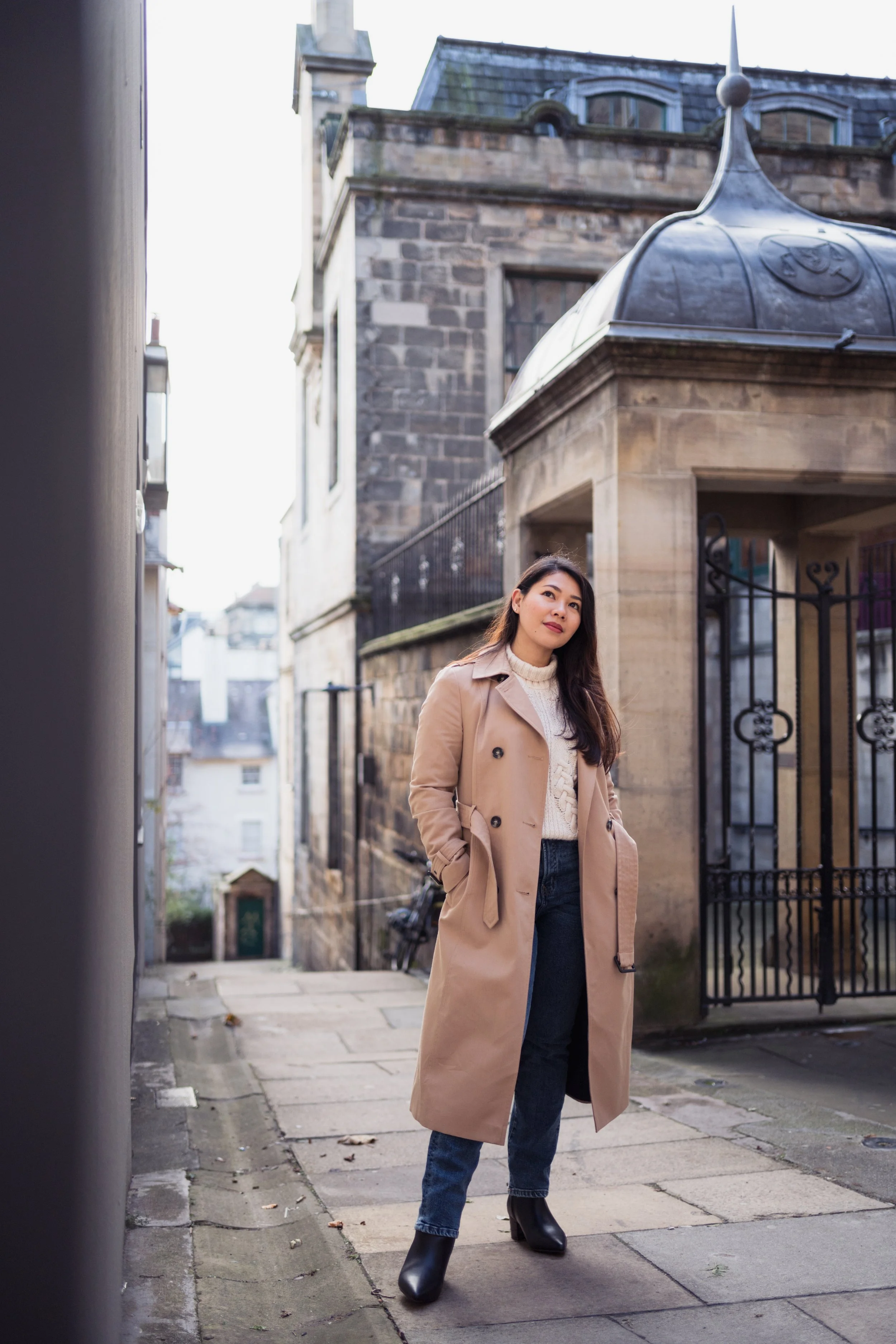 Woman standing on a stone-paved alleyway with historic buildings, wearing a beige trench coat, white sweater, jeans, and black boots.