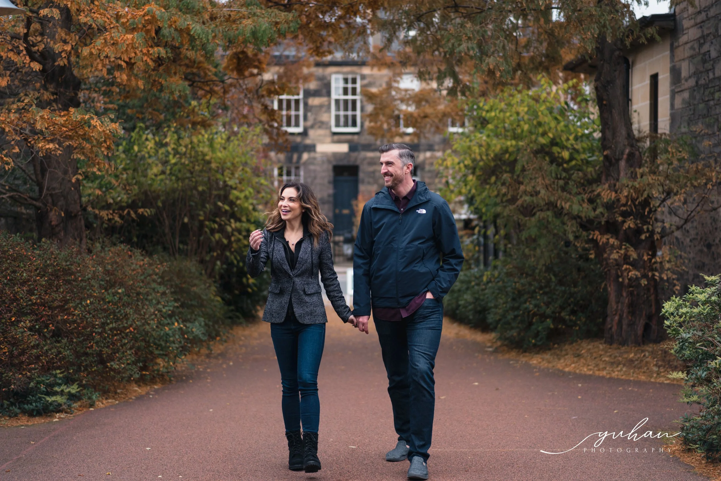 A man and woman walk hand in hand along a tree-lined path with fall foliage, smiling and enjoying a casual outdoor stroll.