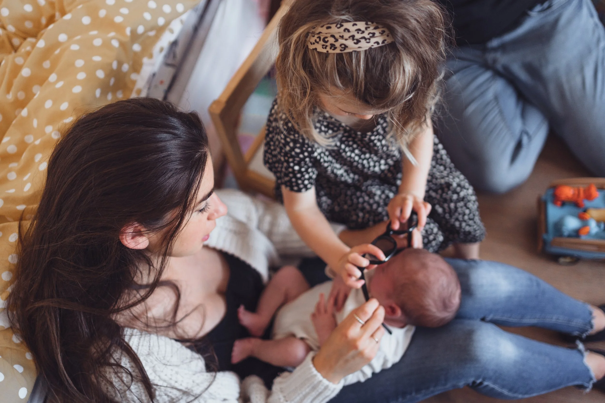 A woman and a young girl sitting on the floor with a baby. The girl is holding a pair of glasses over the baby's face, and the woman is smiling.