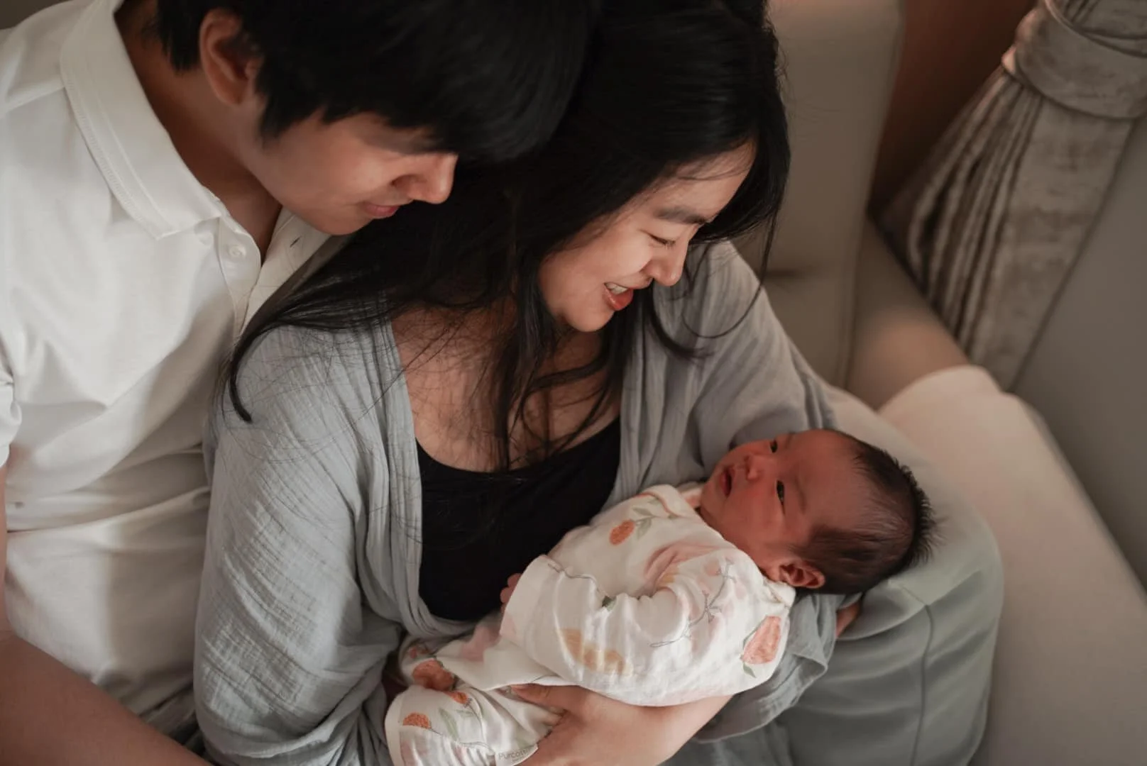 A woman holding a newborn baby while a man leans in, all smiling and looking at each other in a cozy indoor setting.
