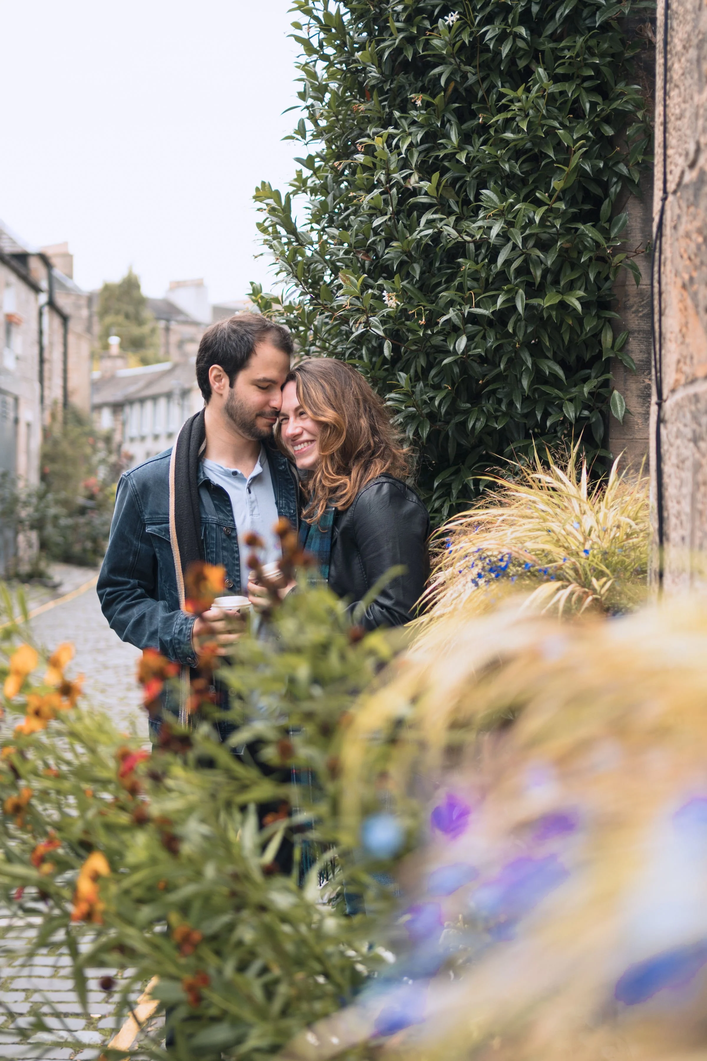 A young couple sharing a tender moment on a city street, standing close to a brick wall decorated with lush green foliage and colorful flowers.