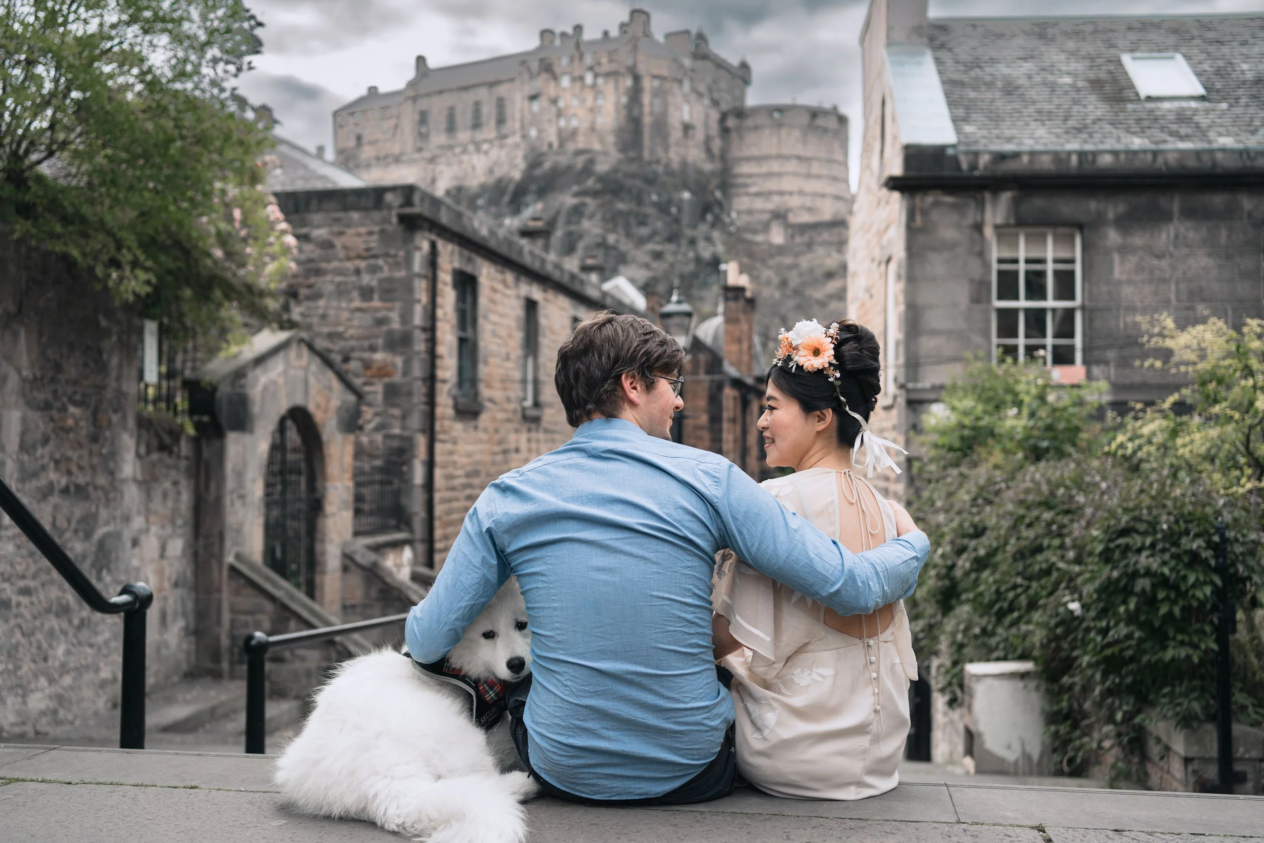 A couple sitting on a stone staircase, with a fluffy white dog beside them, in a historic city with stone buildings and a castle in the background. The woman has a floral hairpiece and they are smiling at each other.