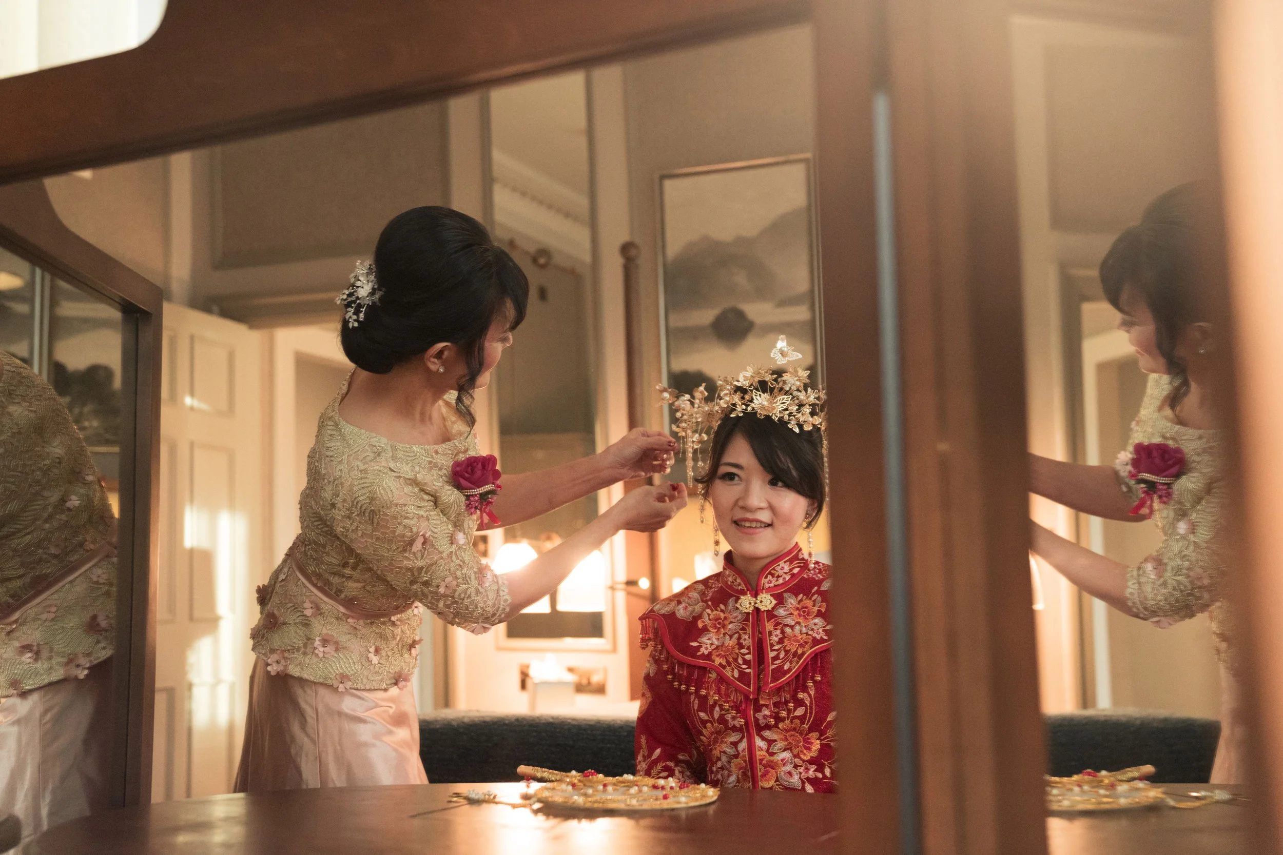 A woman in traditional Chinese wedding attire sitting indoors with an older woman placing a decorative headpiece on her head