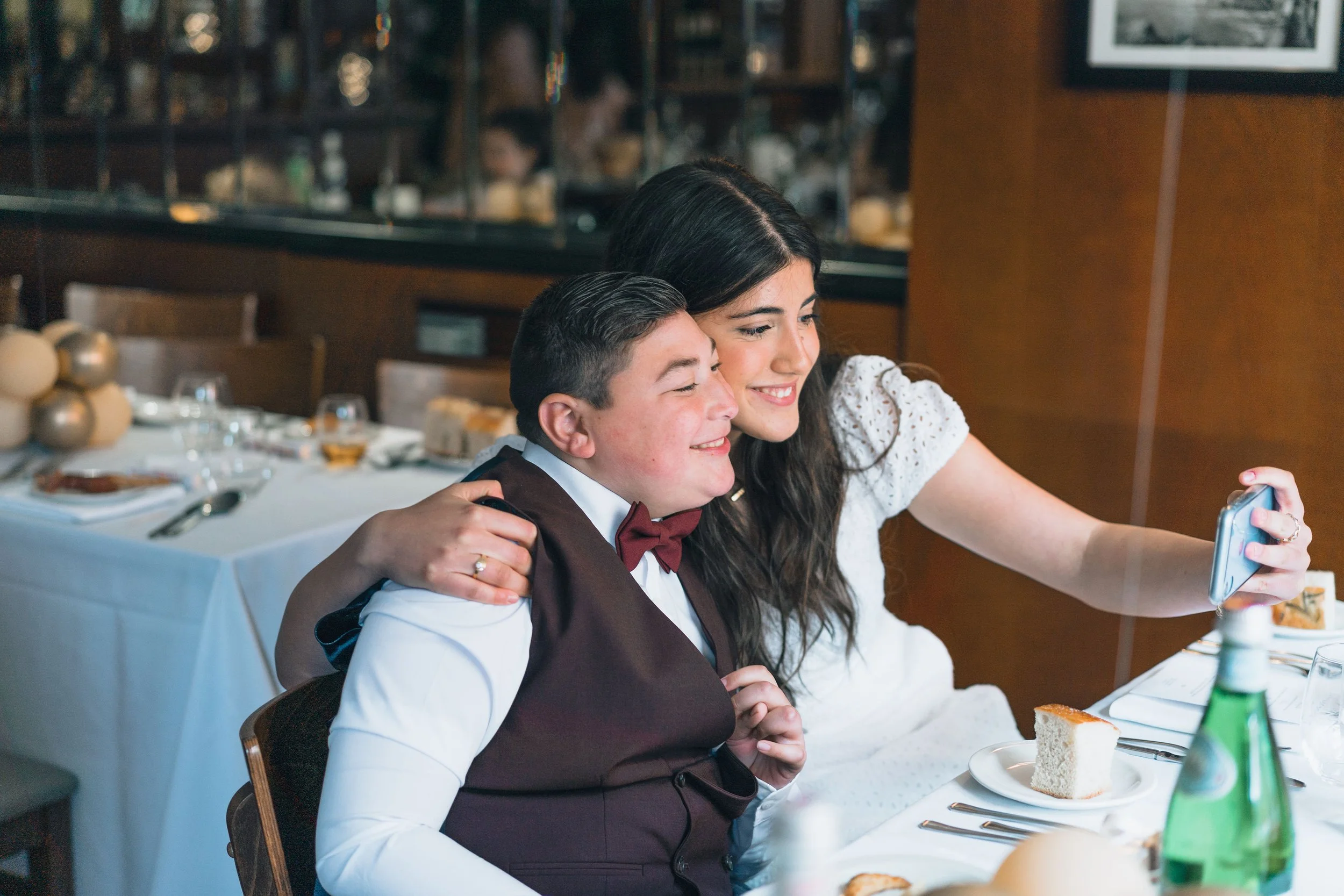 A young woman and a young boy taking a selfie together at a formal dining event. The woman has dark hair and is wearing a white dress, while the boy is dressed in a formal suit with a bow tie. They are smiling happily, seated at a table set with plat