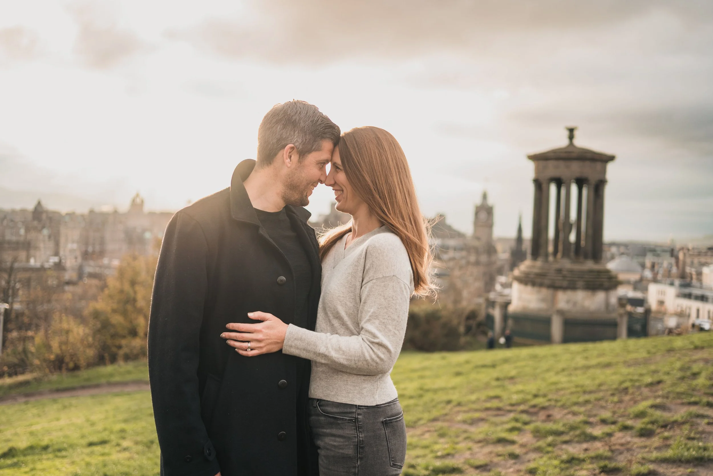 A couple standing close together outdoors, touching foreheads and smiling, with a cityscape and ruins in the background during sunset.