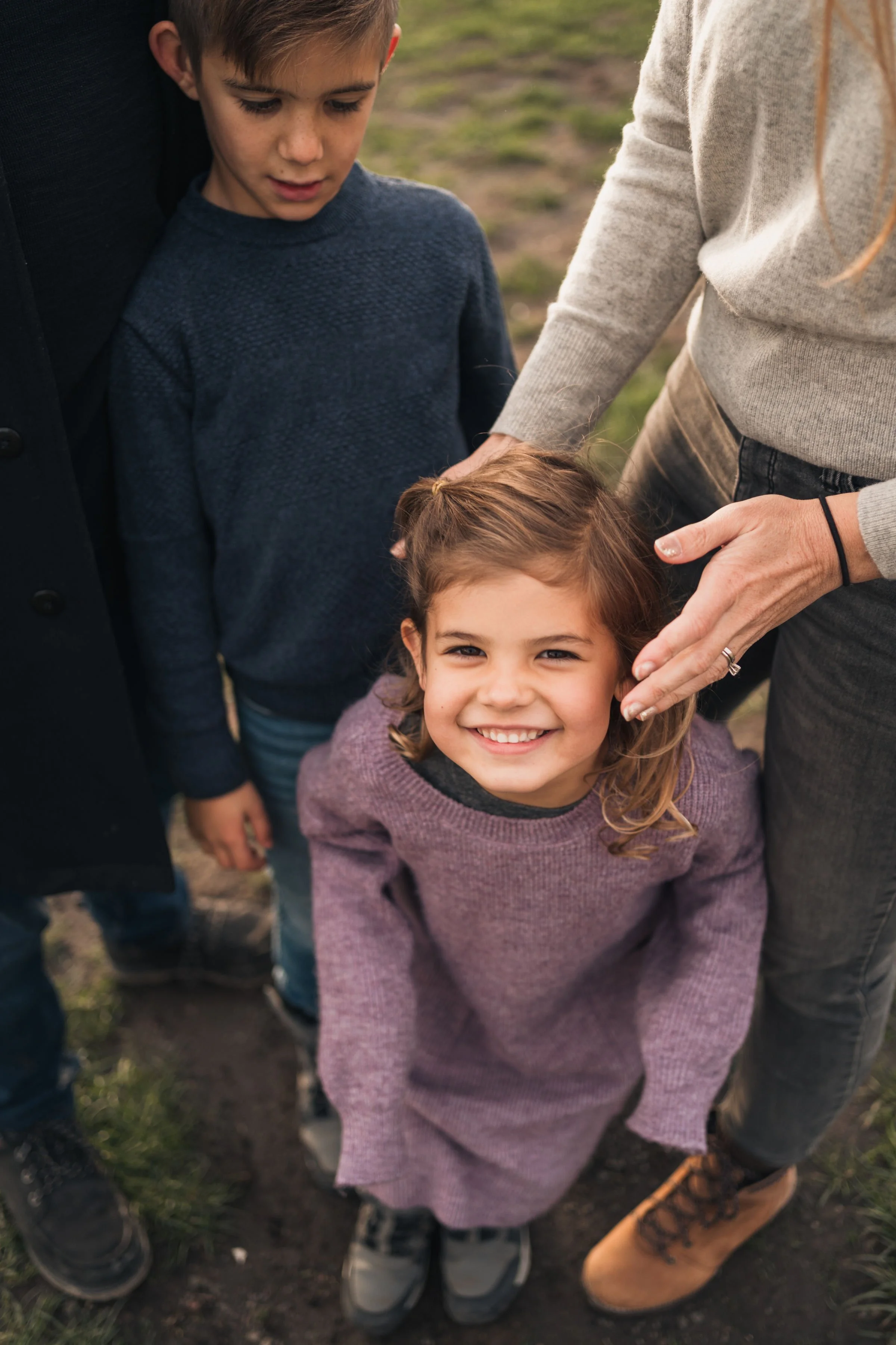 A smiling young girl standing outdoors on dirt, with two children and two adults around her, all dressed in warm clothing.