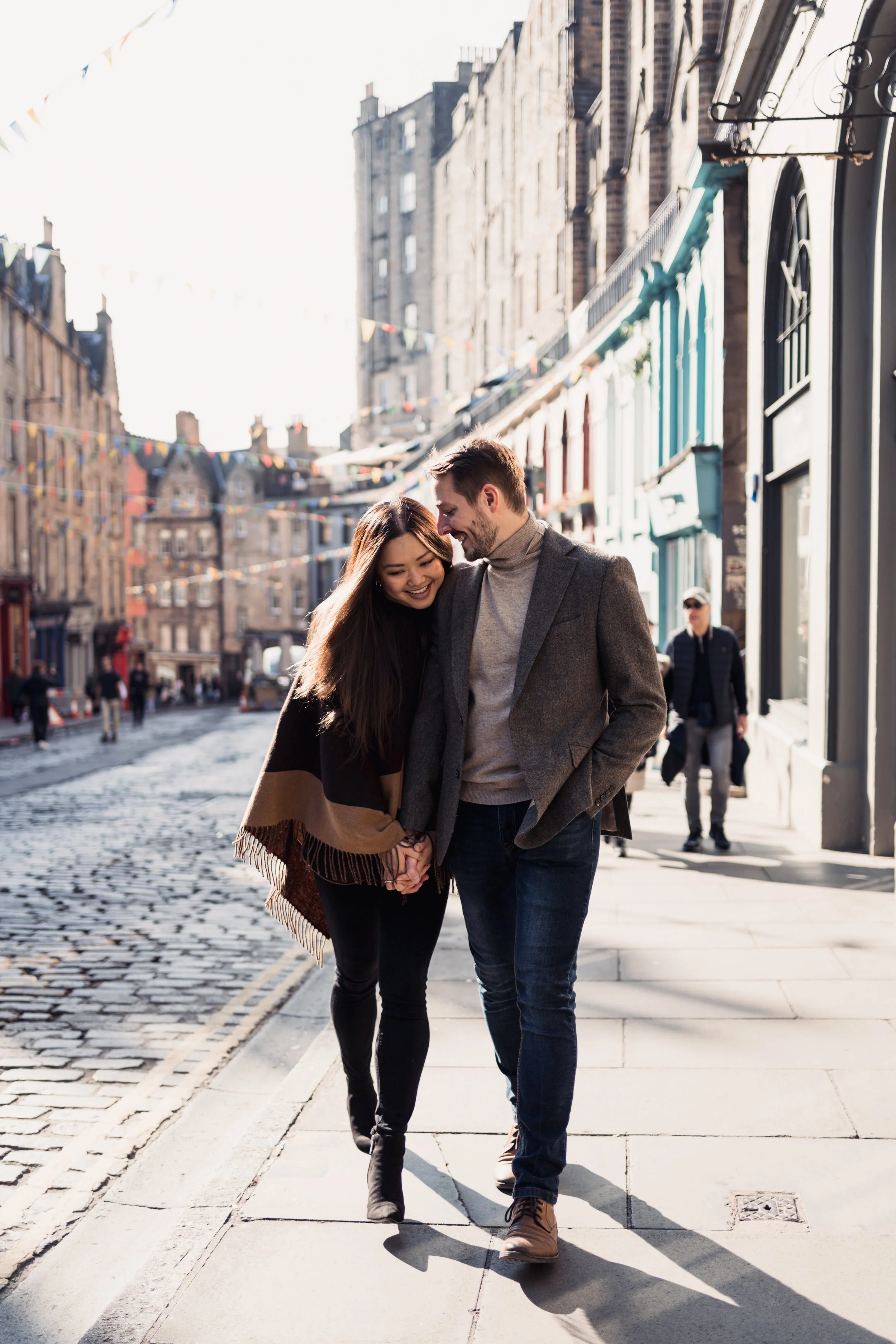 A young couple walking close together on a city sidewalk, holding hands and smiling, with colorful buildings and street decorations in the background.