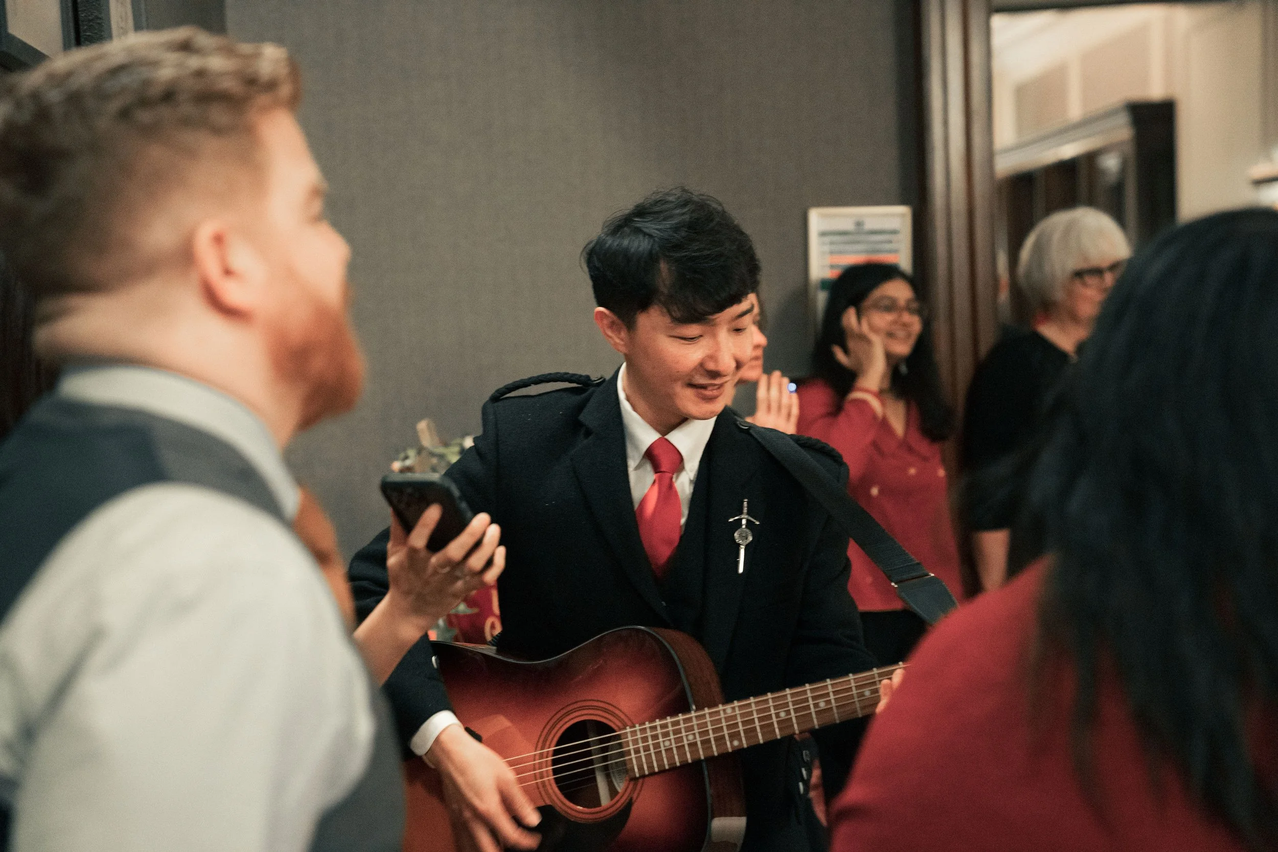 Young man in formal attire playing guitar at social gathering, surrounded by smiling people.