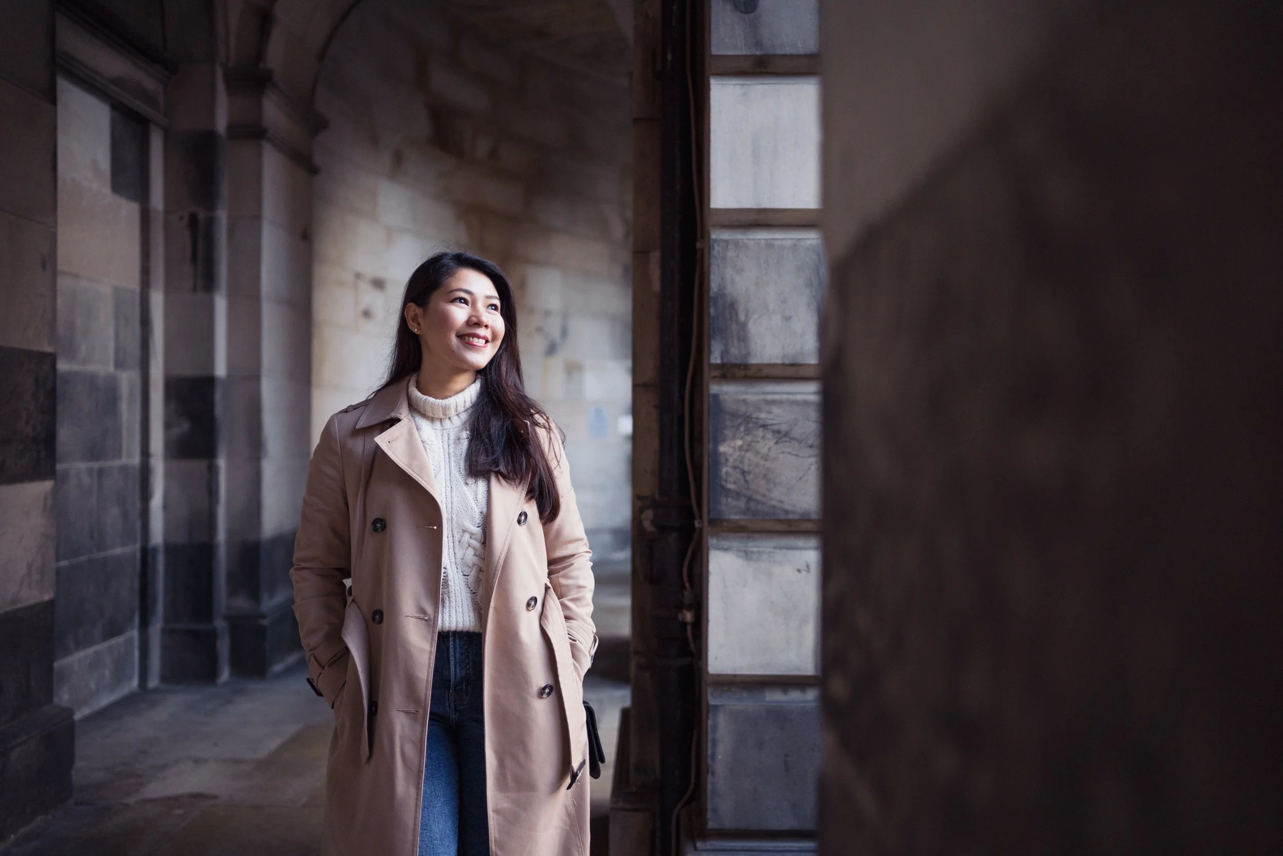 A young woman with long dark hair smiling and standing with her hands in the pockets of a beige trench coat, in an outdoor stone archway.