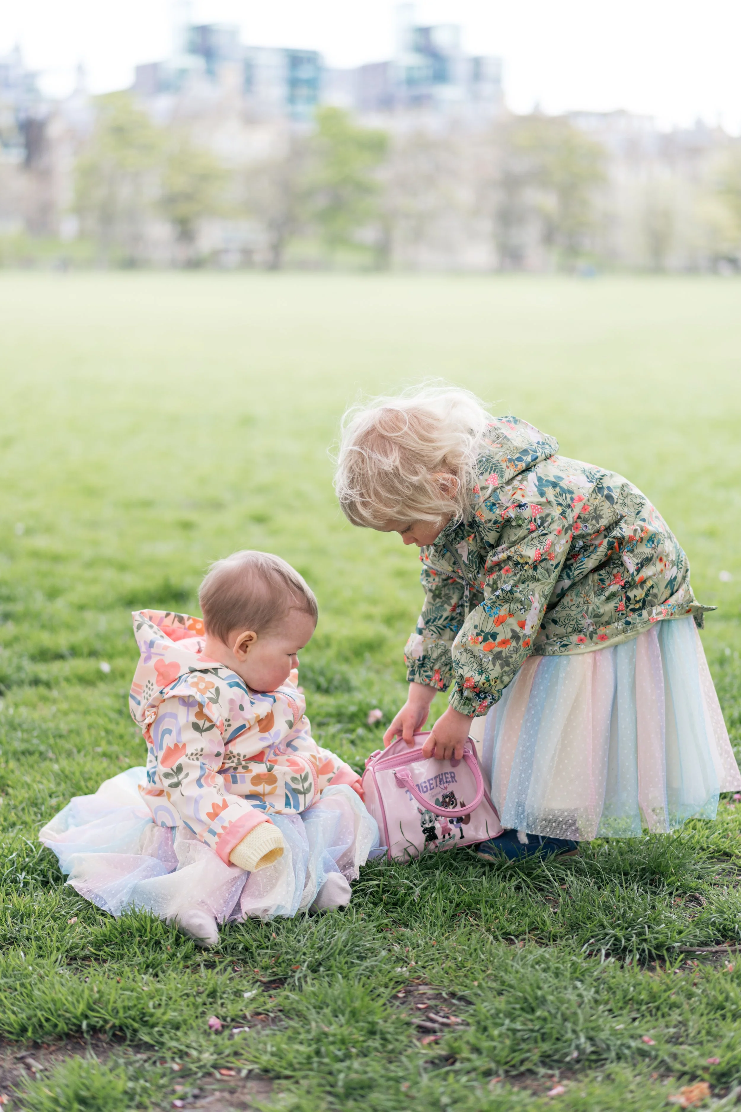 Two young children, one sitting on the grass and the other standing, are playing together in a park. They are dressed in colorful, patterned jackets and skirts, with one child holding a pink bag. The background shows green grass, trees, and blurred b