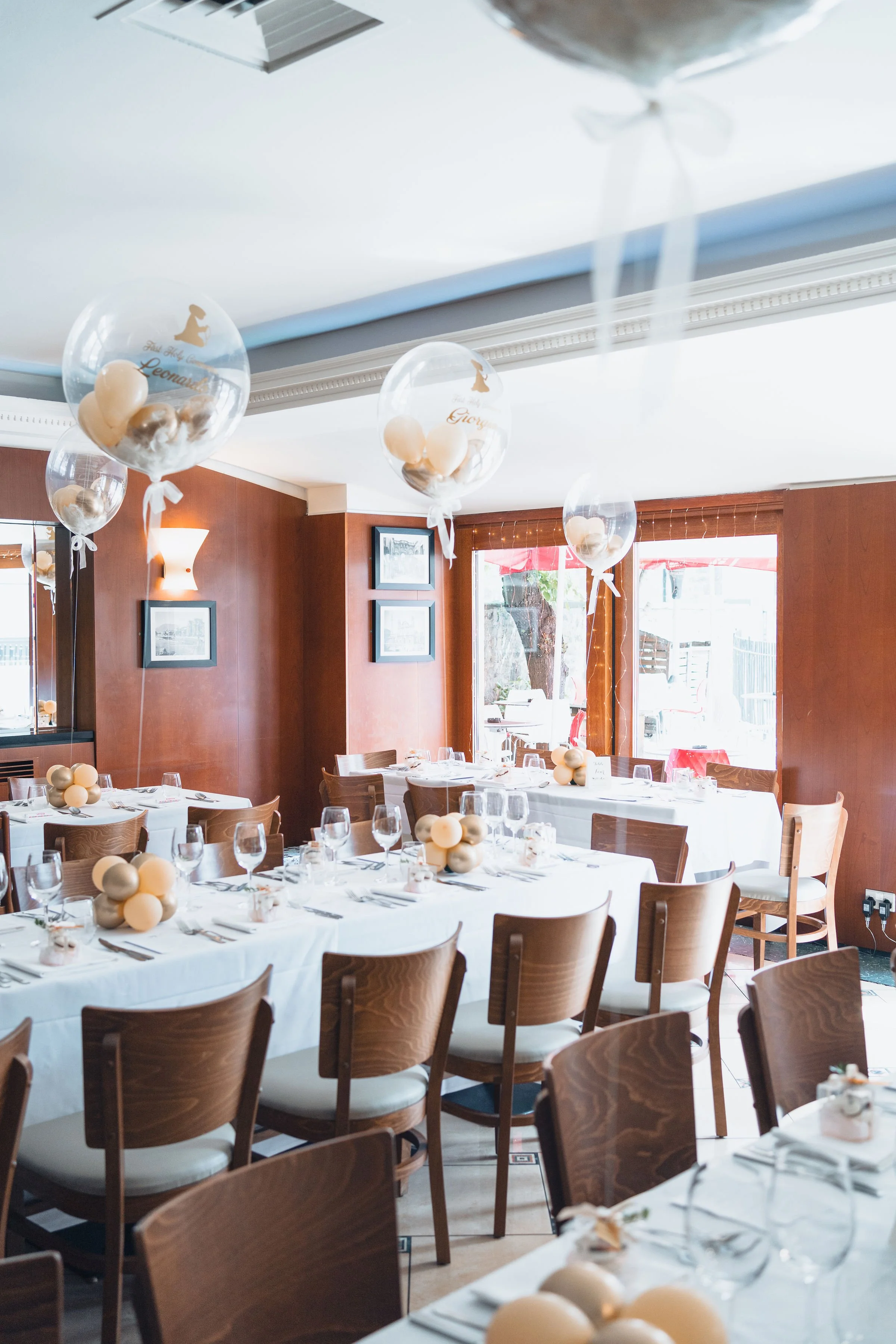 Decorated dining room with white tablecloths, wooden chairs, and balloon centerpieces, set up for an event with plates, glasses, and utensils.
