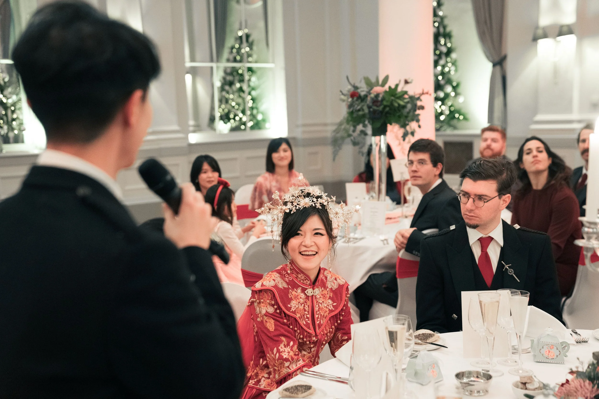 A woman in traditional red Chinese attire and a floral headpiece is smiling at a man with a microphone, during a celebration at a decorated banquet hall with Christmas trees and floral arrangements.