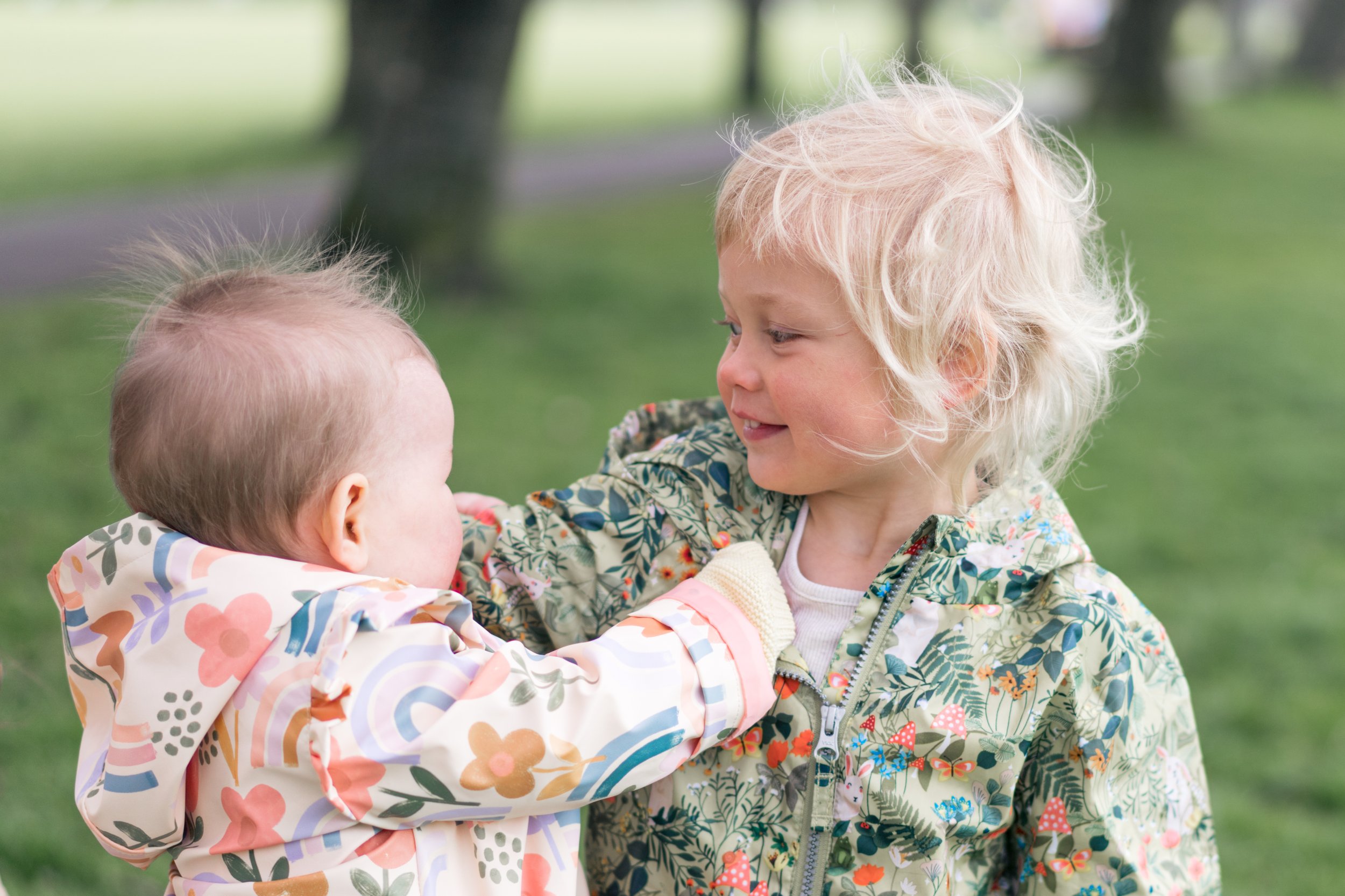 Two young children, one with blonde hair and one with light brown hair, are playing together outdoors on a grassy area with trees in the background. The blonde girl is smiling and looking at the other child, who is touching her face.