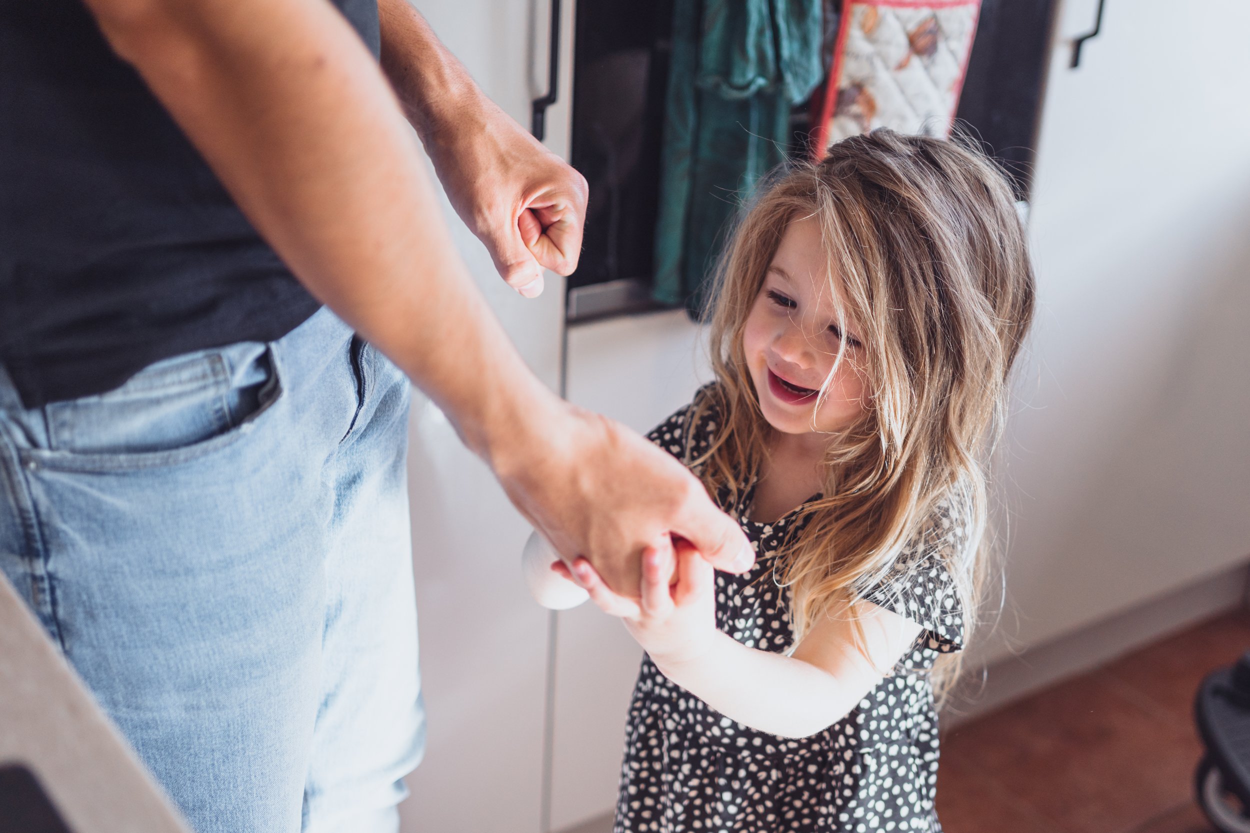 A young girl with long, wavy hair and a black and white polka dot shirt is smiling and holding an object while an adult, wearing a black shirt and blue jeans, is showing her something with their hands in a kitchen.