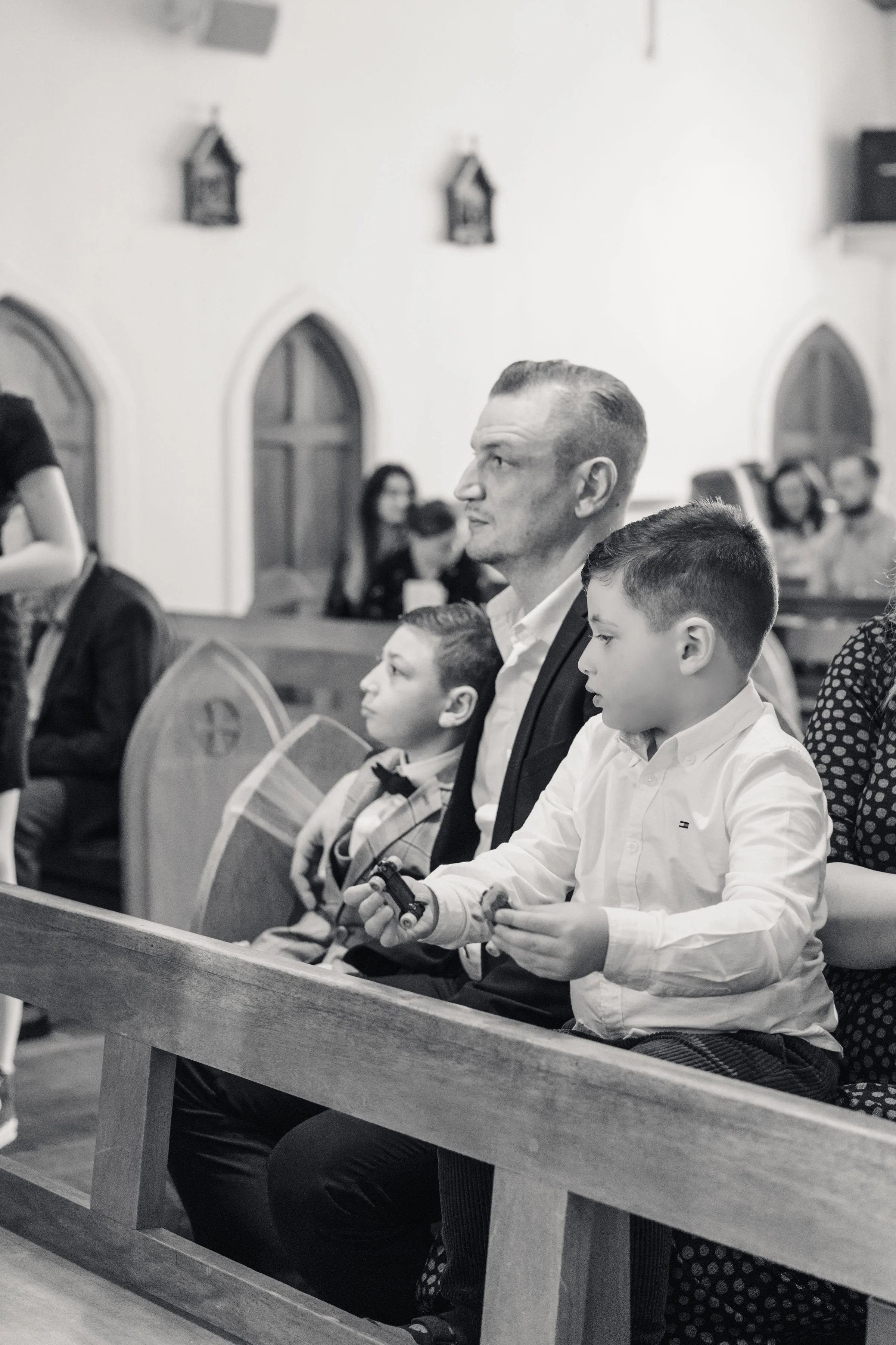 A black and white photo of people sitting in a church. A man and two boys are seated in the front row, with the boys holding toys.