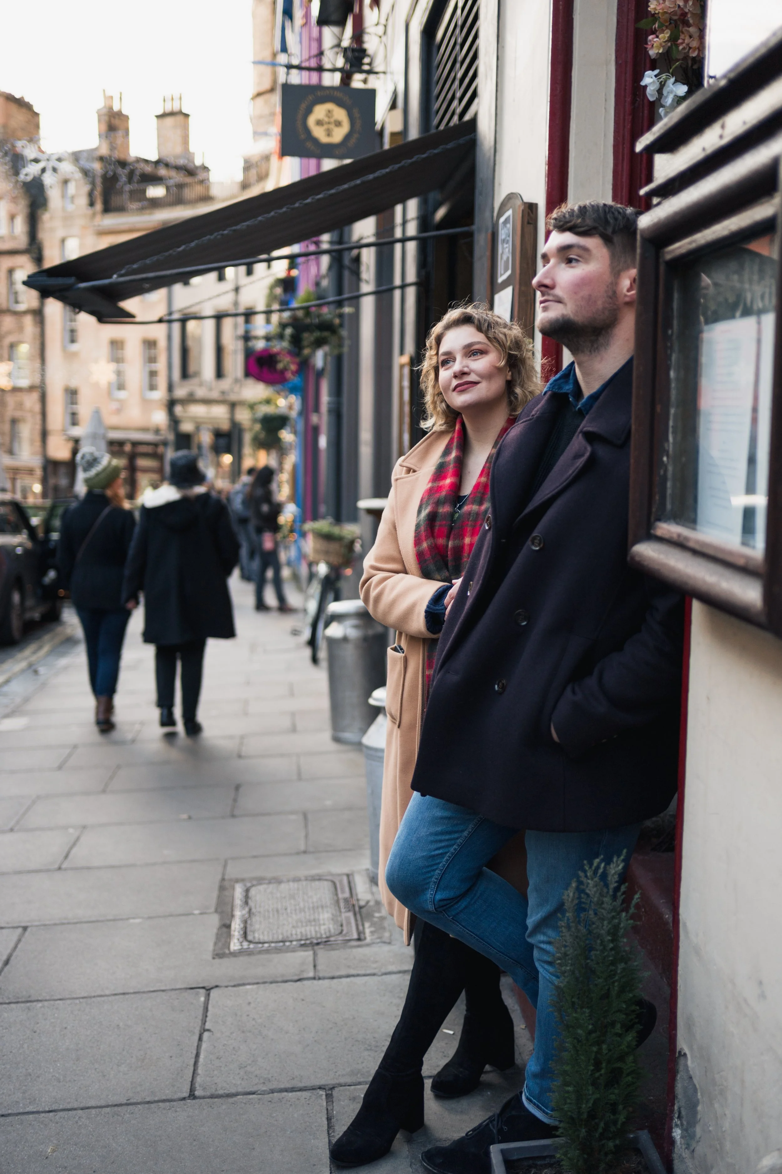A young man and woman standing outside a storefront on a city street, leaning against the wall, with other pedestrians walking by in the background.