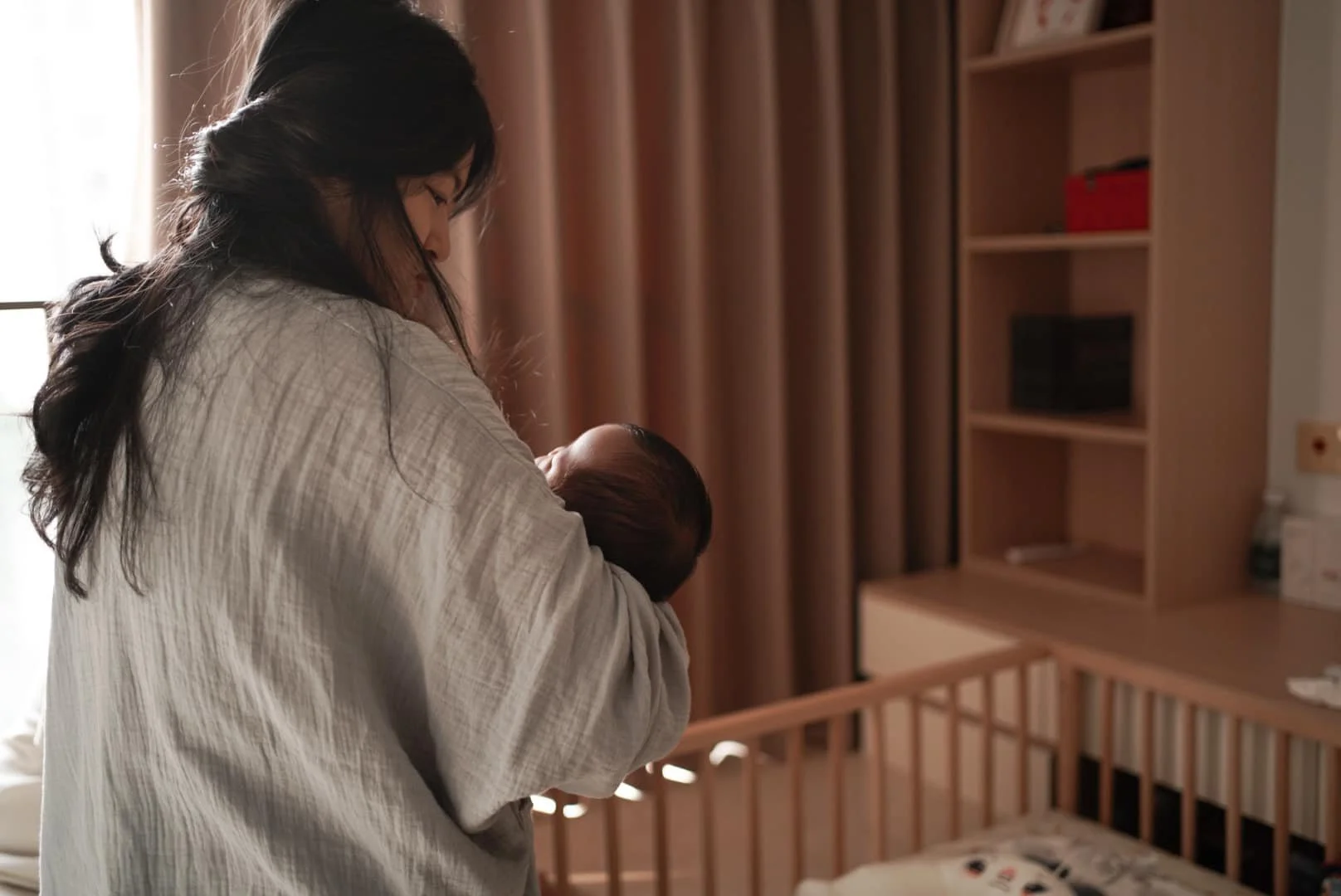 Woman holding a newborn baby in a nursery with wooden furniture and shelves.