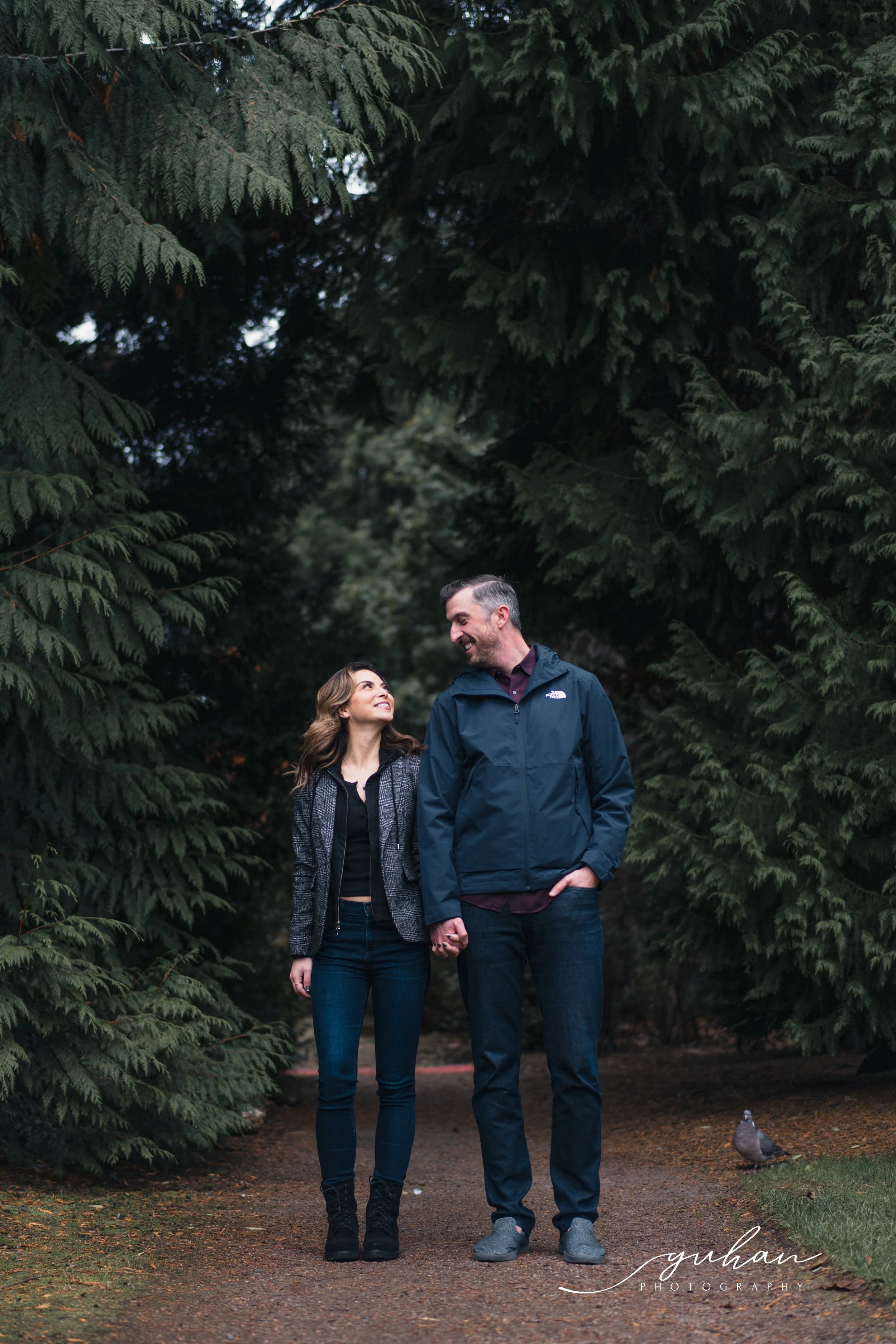 A couple holding hands and smiling at each other walking on a forest path surrounded by tall green trees, with a pigeon on the ground nearby.