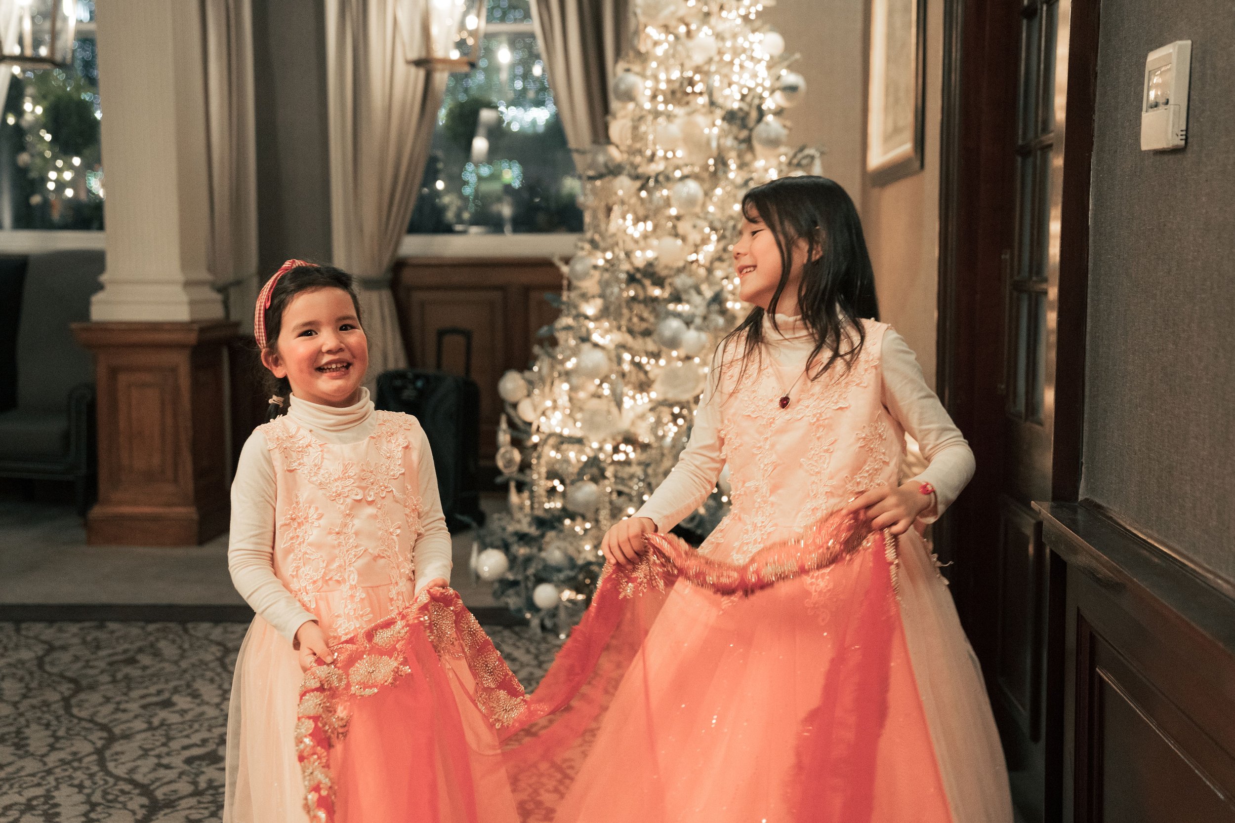 Two young girls in peach-colored dresses with lace details, smiling and playing with a fabric material, in front of a decorated white Christmas tree with ornaments and lights, indoors at night.