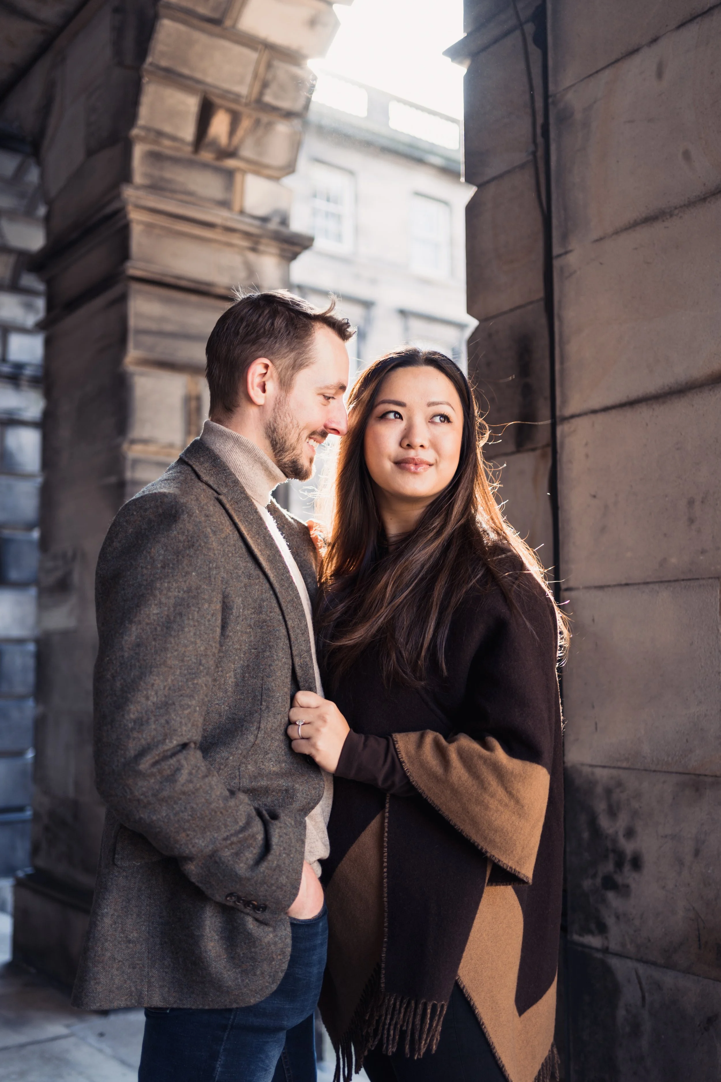 A couple standing close together outside near a stone wall, with the man smiling and the woman gazing thoughtfully into the distance.