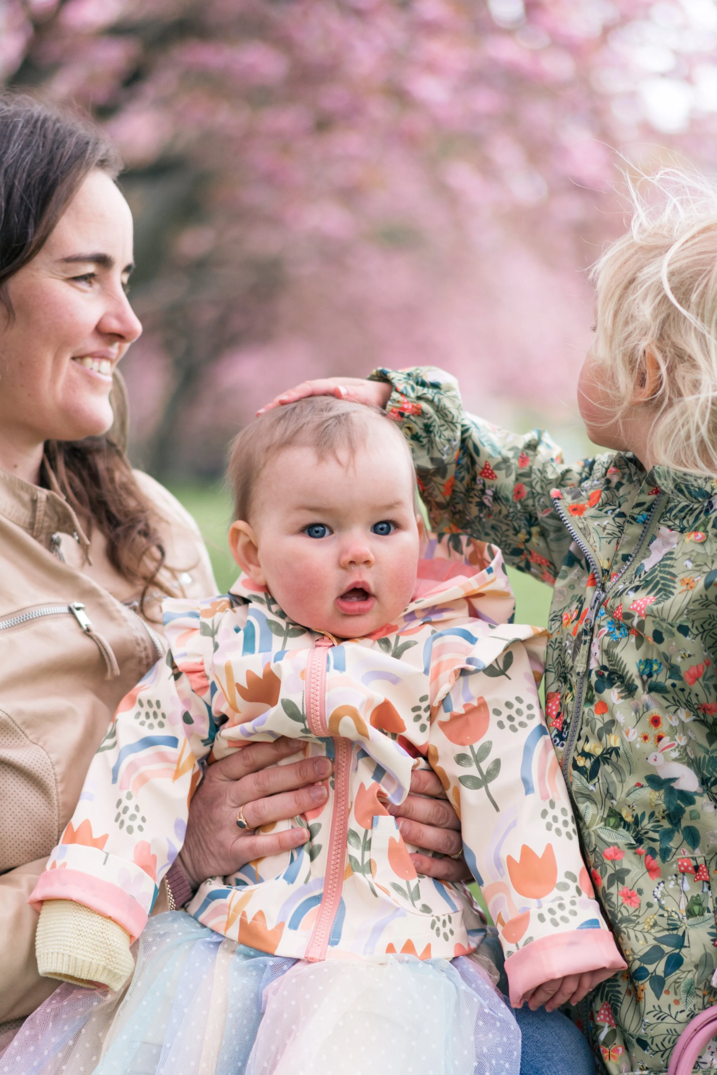 A woman holding a surprised looking baby girl with blue eyes and a floral jacket, while another child pets the baby's head, with pink flowering trees in the background.