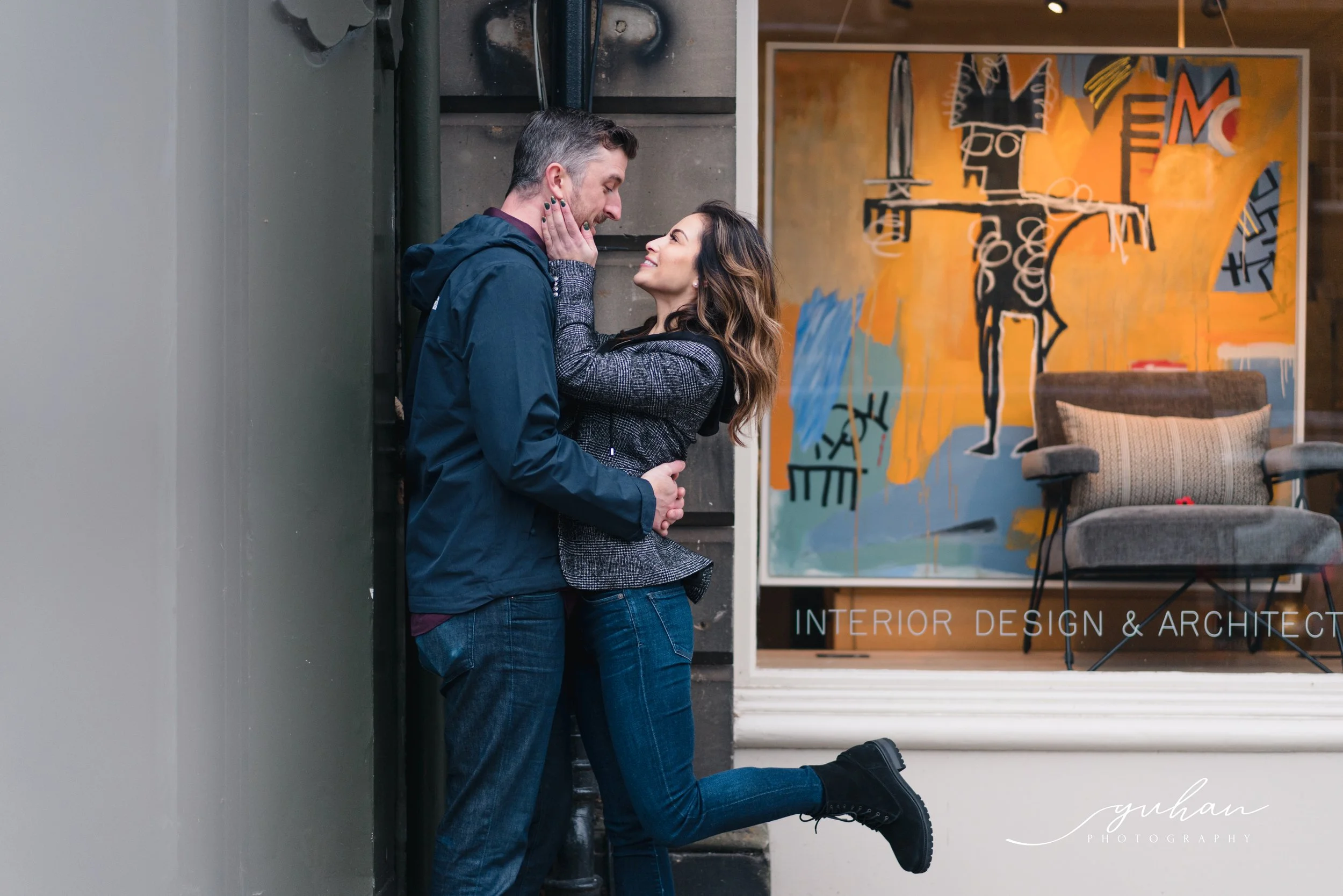 A couple faces each other closely in front of an interior design store window, with the woman's right leg raised. The window features colorful abstract art and the text 'INTERIOR DESIGN & ARCHITECT'.