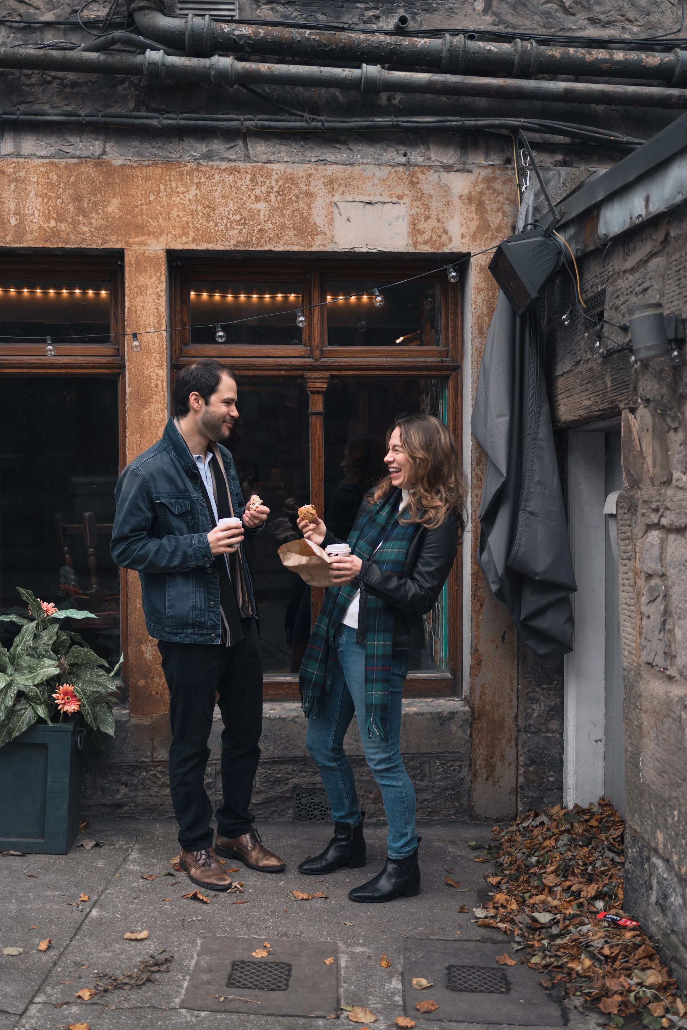 A man and woman laugh and enjoy snack foods outdoors in a narrow alleyway at night, behind a window with string lights, on a fall evening.