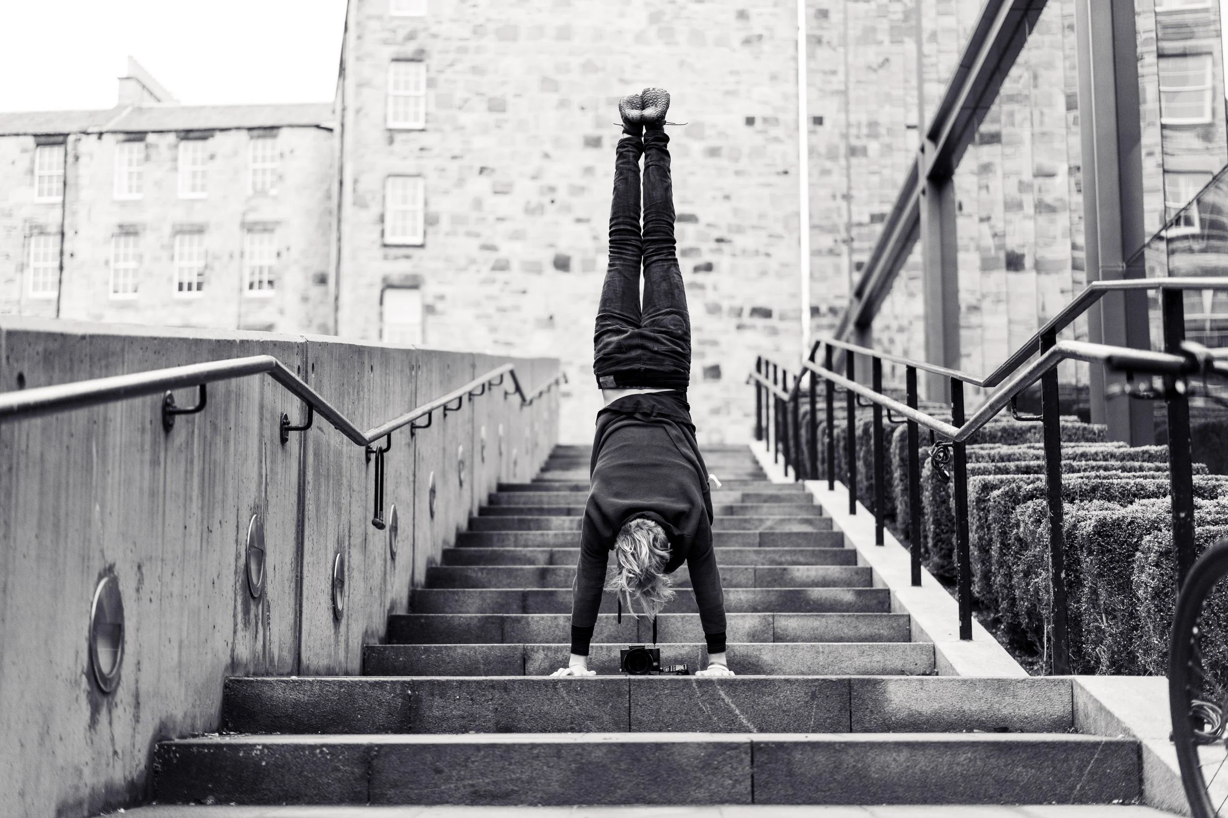 Person performing a handstand on outdoor stairs with a camera placed on the steps, city buildings in the background.