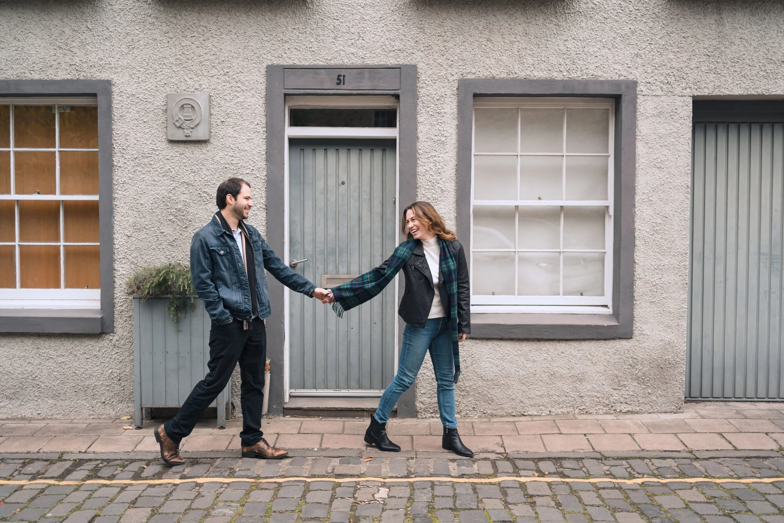A man and woman holding hands and smiling while walking on a cobblestone sidewalk in front of a beige stucco building with two windows and a door.