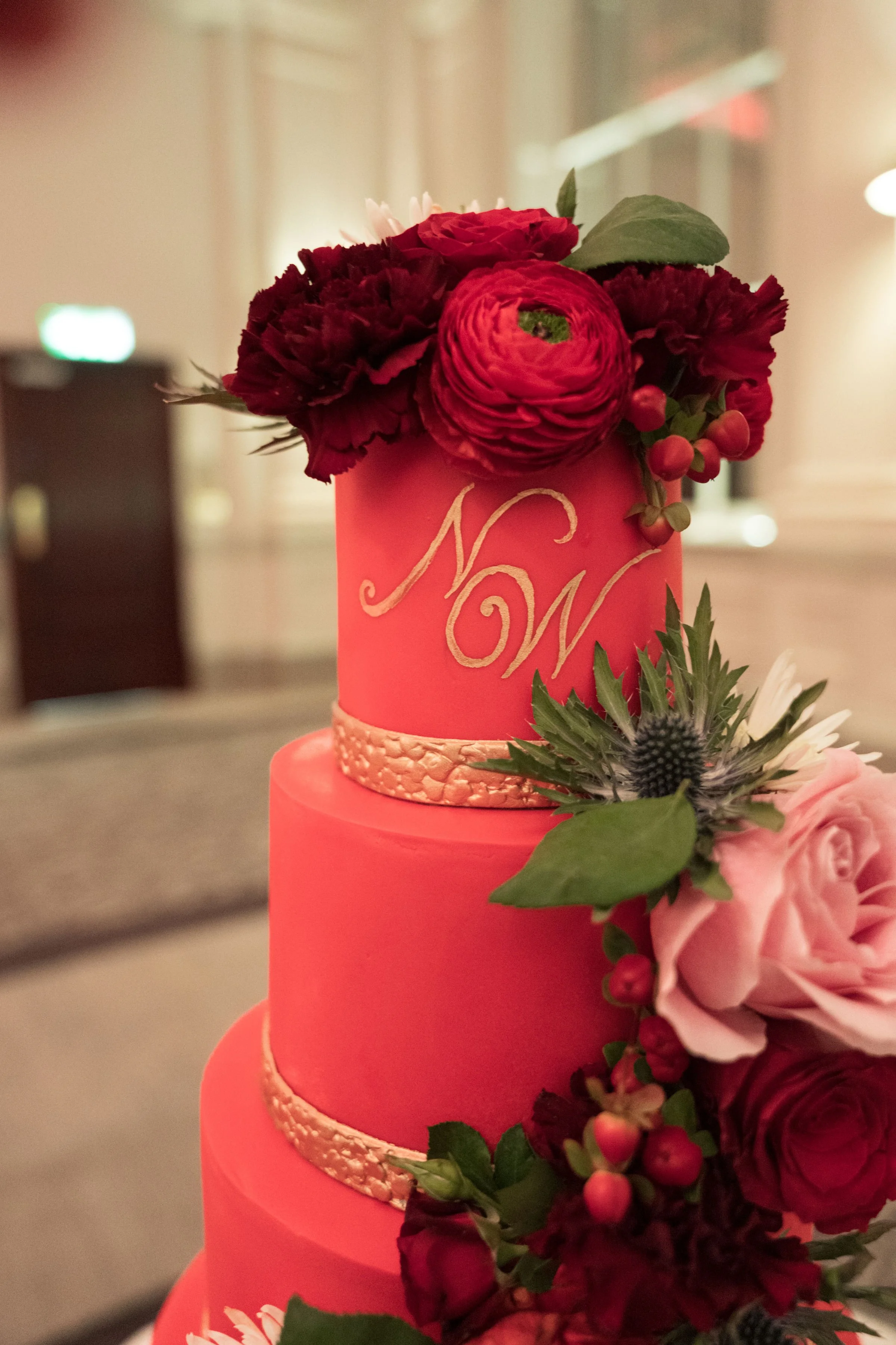 A pink, three-tiered wedding cake decorated with red and pink flowers, green leaves, and a white script indicating the initials "N" and "W" on the middle tier.