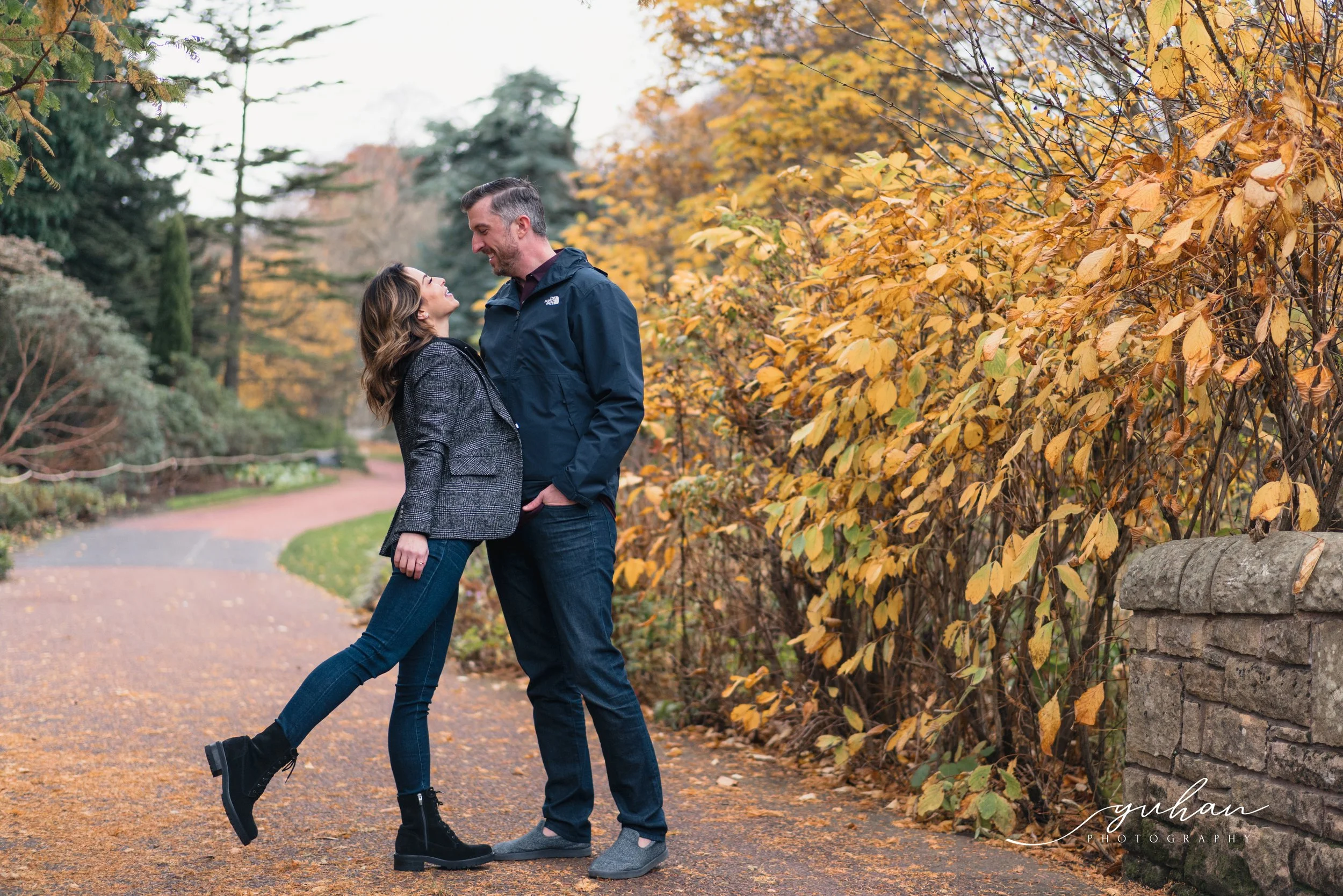 A couple standing on a paved park path surrounded by fall-colored trees and bushes, smiling and looking into each other's eyes.