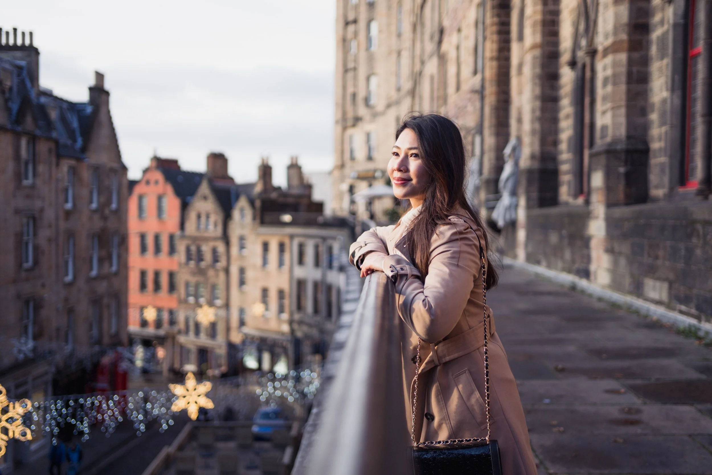 A woman in a beige trench coat leaning on a railing on a balcony, looking to the right with a city street with old buildings and Christmas decorations in the background.
