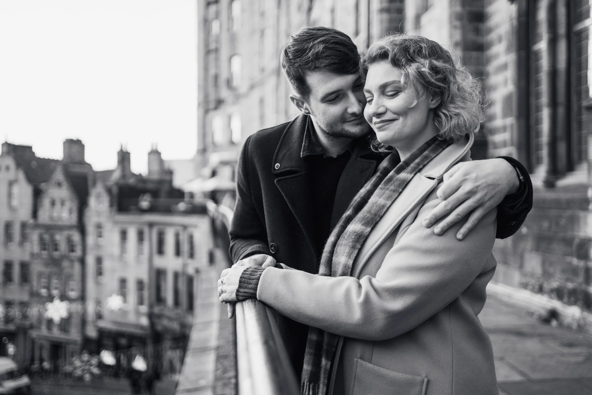 A black-and-white photo of a couple embracing outdoors on a city street, with the man leaning his head against the woman's forehead and smiling, while the woman is smiling with her eyes closed. Buildings are visible in the background.