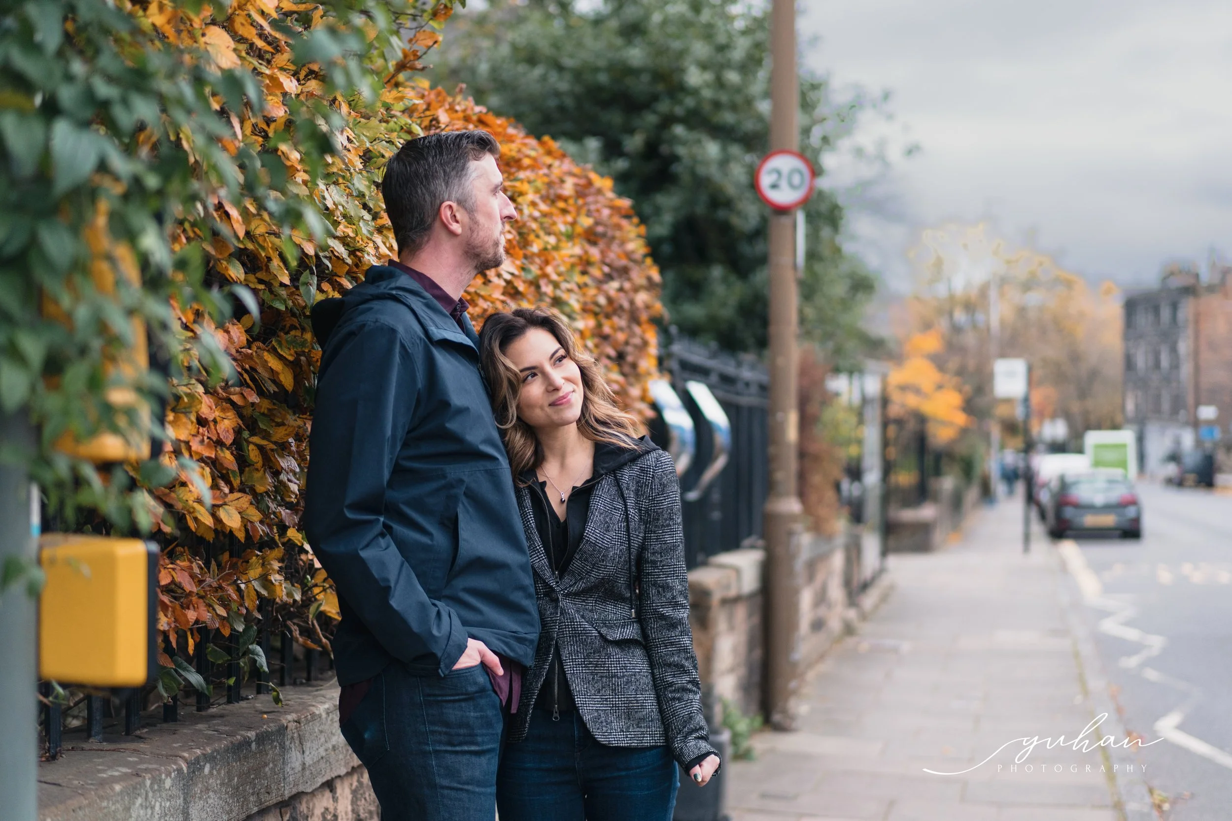A man and woman standing on a sidewalk next to a hedge with autumn leaves, looking into the distance.