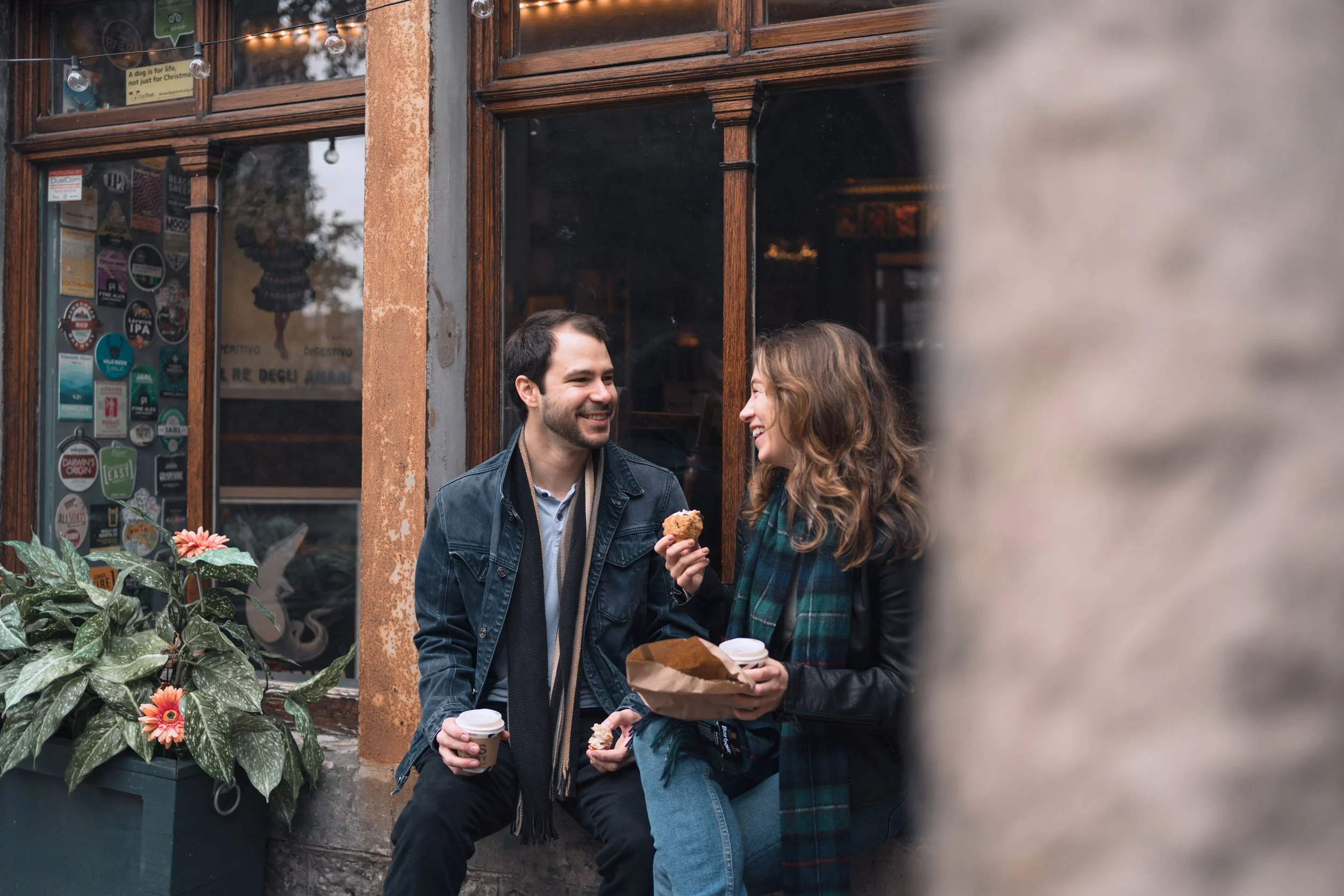 A man and woman sitting outside a café, smiling at each other. The woman is holding a coffee cup and a pastry, while the man also has a coffee cup. They appear to be enjoying a casual moment together near a window with stickers and a plant with pink 
